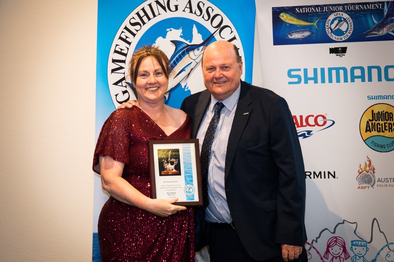 A woman in a red dress holding a framed certificate, standing next to a man in a suit at a fishing award event, with banners of the GamEing Fishing Association and other fishing sponsors behind them.