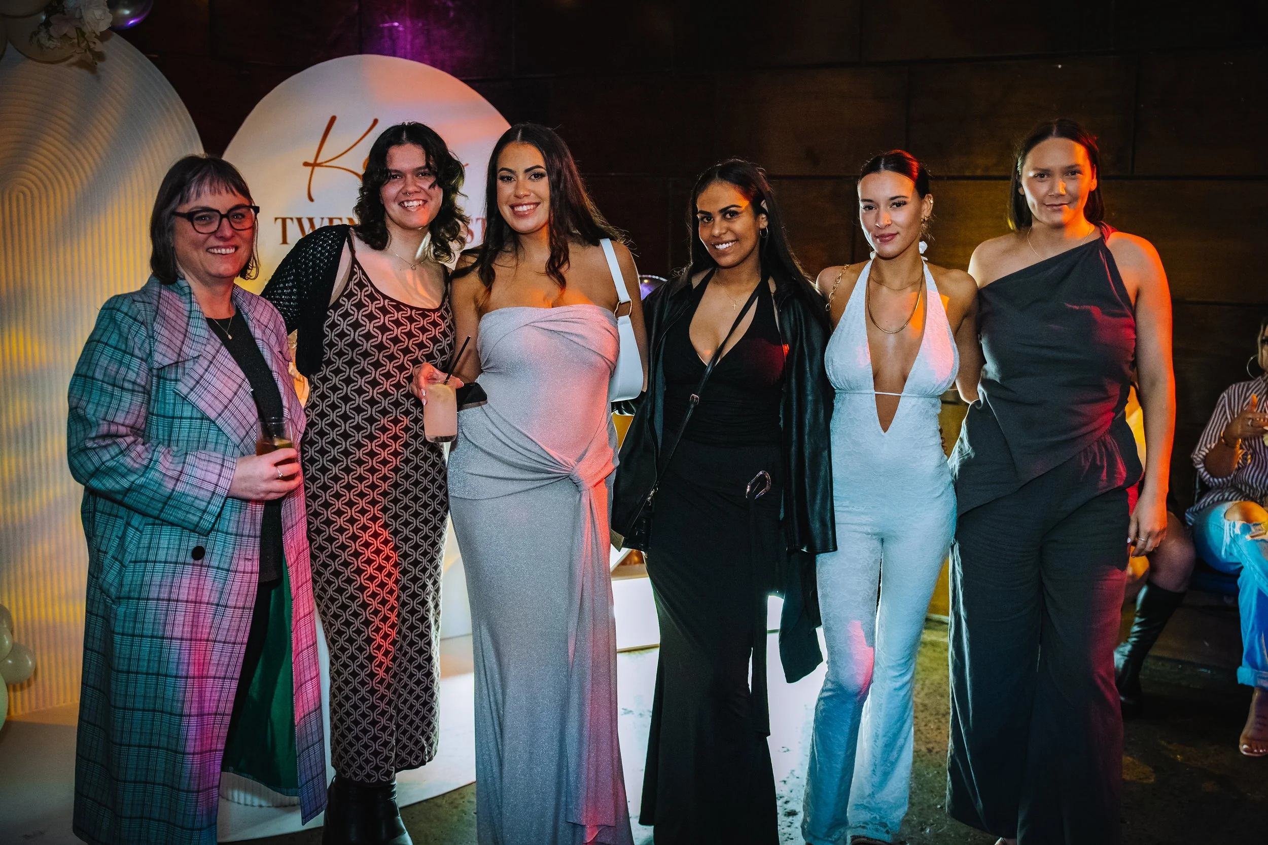 Group of seven women standing together at an indoor event, smiling at the camera, with some holding drinks, in front of a backdrop with a circular sign that says 'K TW...'