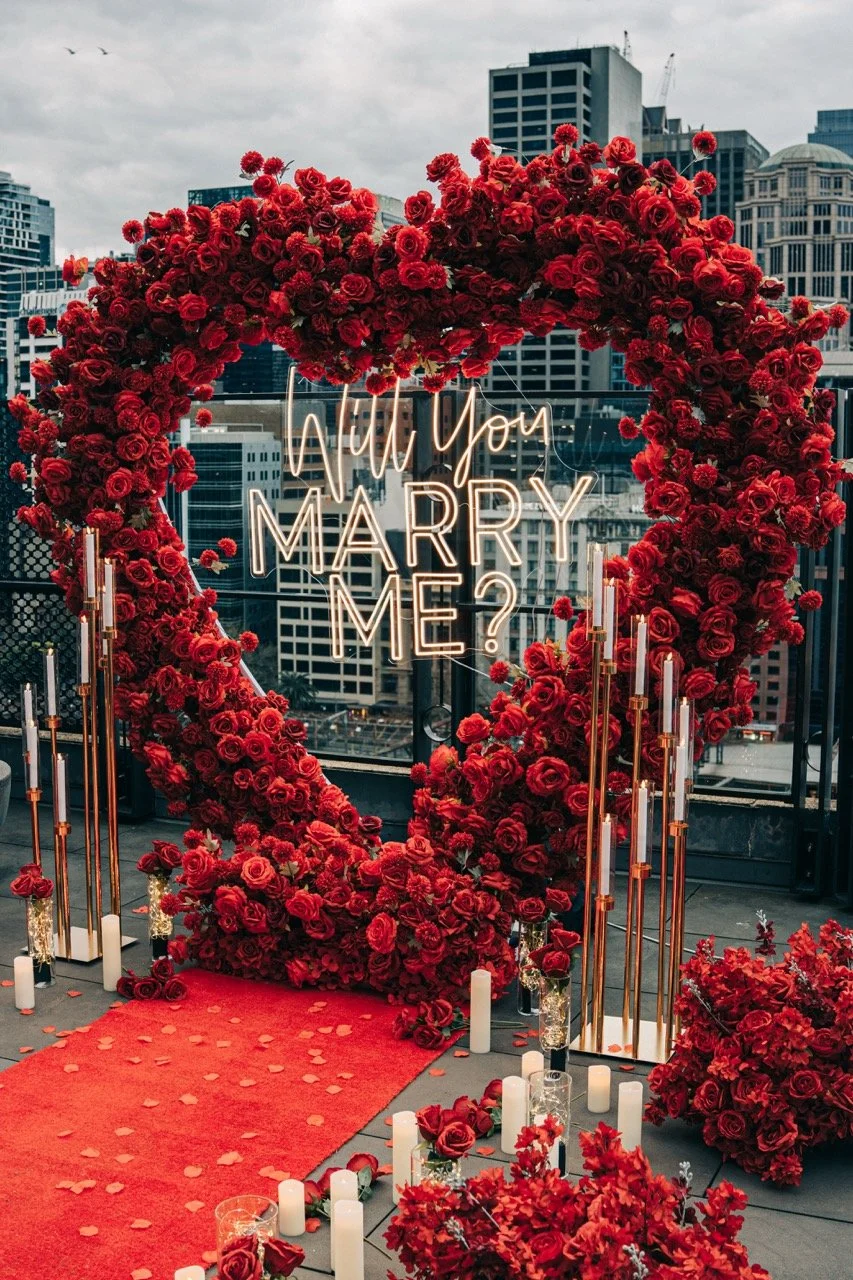 A romantic rooftop setup with a large red roses heart arch and a neon sign that says "Will You Marry Me?" surrounded by candles and floral arrangements, set against a city skyline.