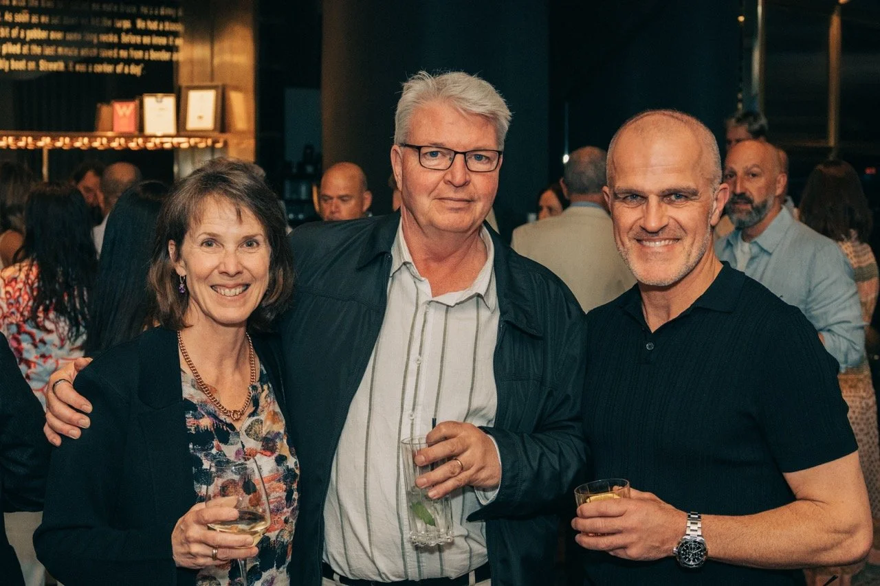 Three people smiling and holding drinks at a social event, with a crowd in the background.