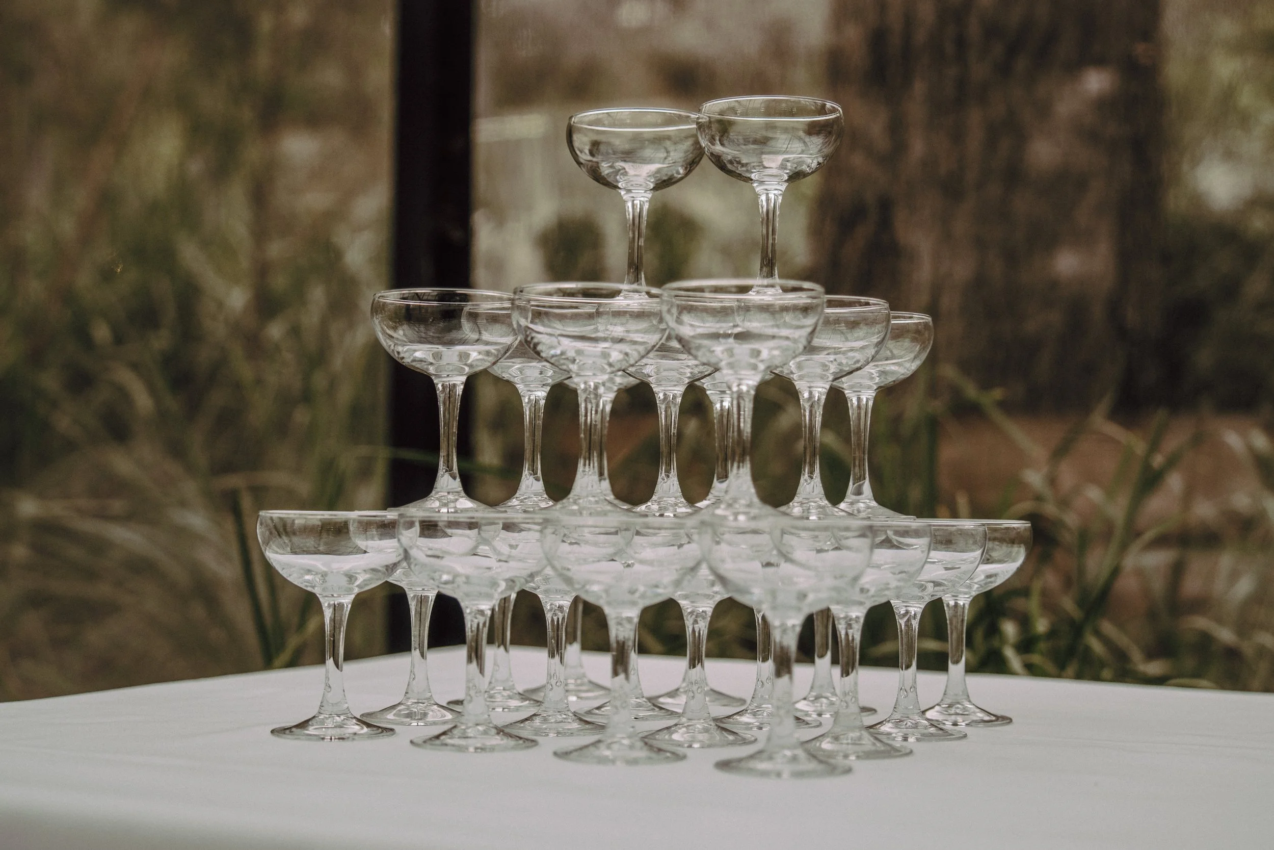 A pyramid of empty champagne glasses stacked on a white table with a blurred outdoor background.
