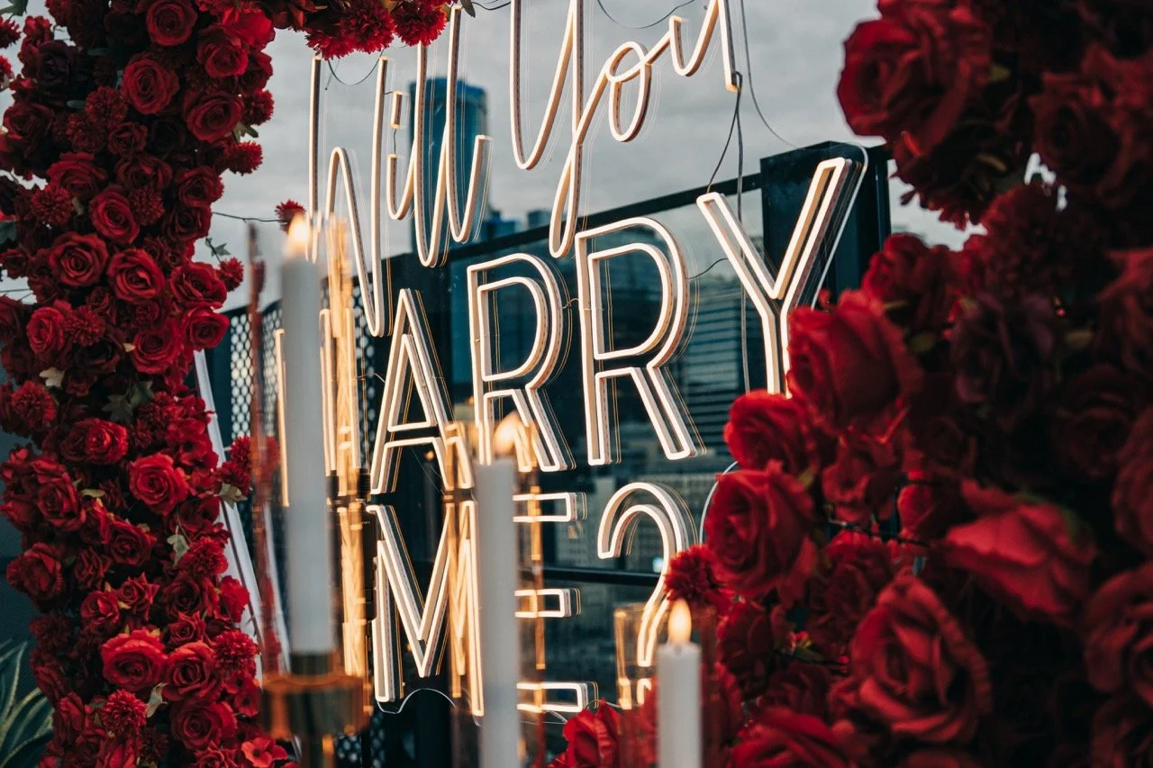 Neon sign reading 'Will you marry me?' surrounded by red roses and candles.