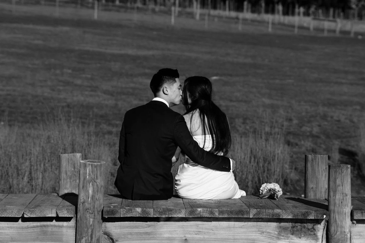 A black and white photo of a couple sitting close together on a wooden bench in a field near a line of trees, about to kiss.