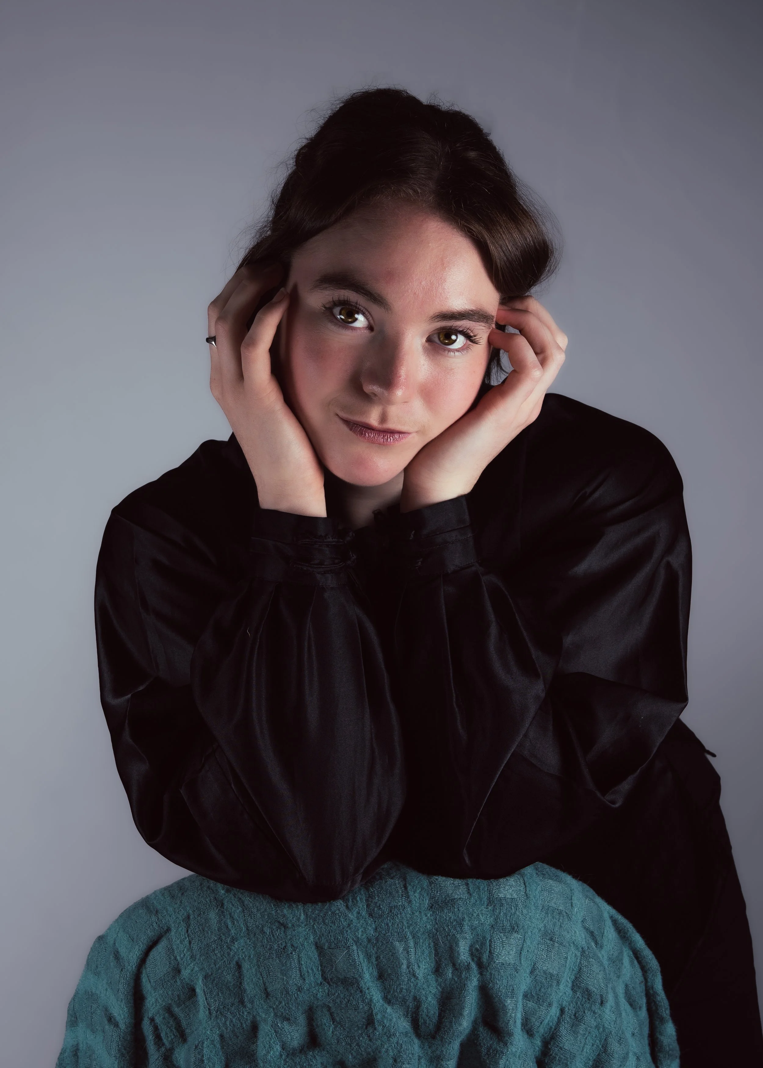 A young woman with dark brown hair, green eyes, and a fair complexion, wearing a black satin shirt, posed with her hands touching her face, seated on a teal textured chair, with a neutral gray background.