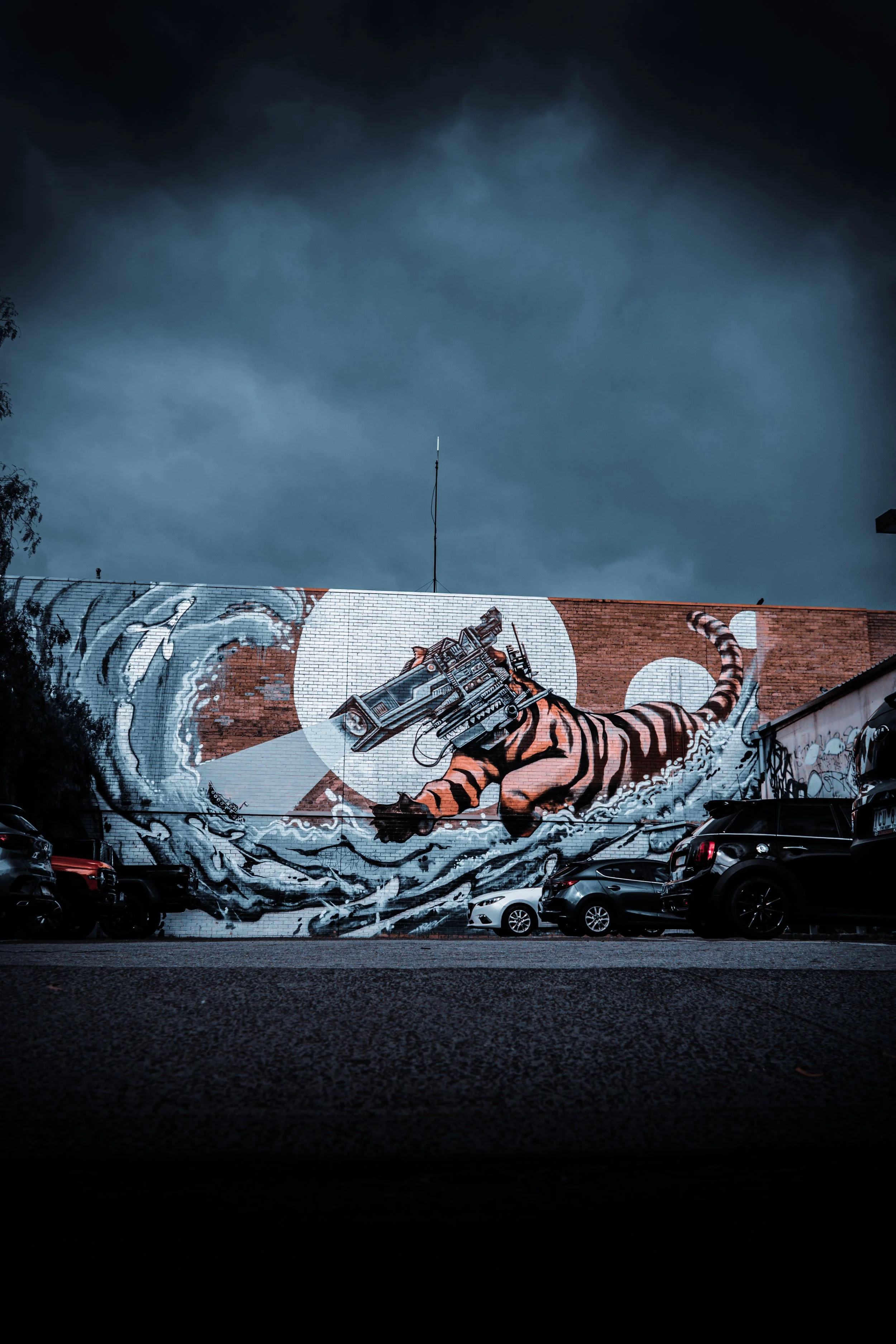 Street art mural of a tiger with a mechanical tiger head, surrounded by water and waves, painted on a brick wall during cloudy weather, with parked cars in front.