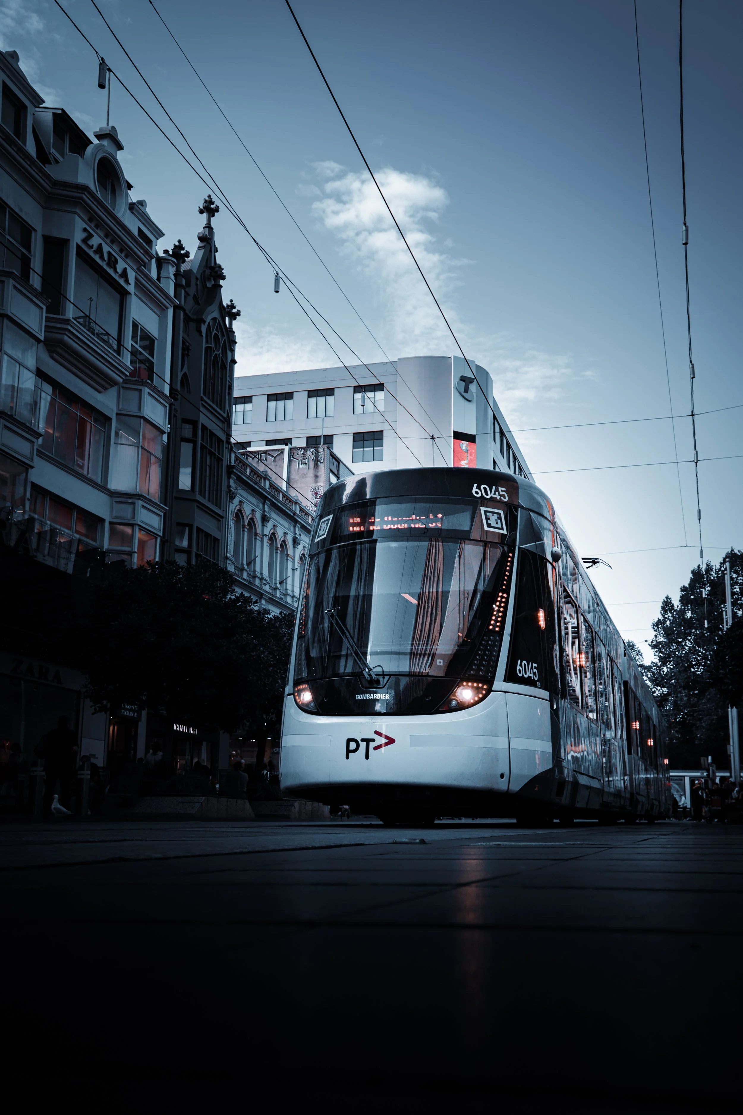 Modern tram on city street with buildings and overhead wires during dusk.