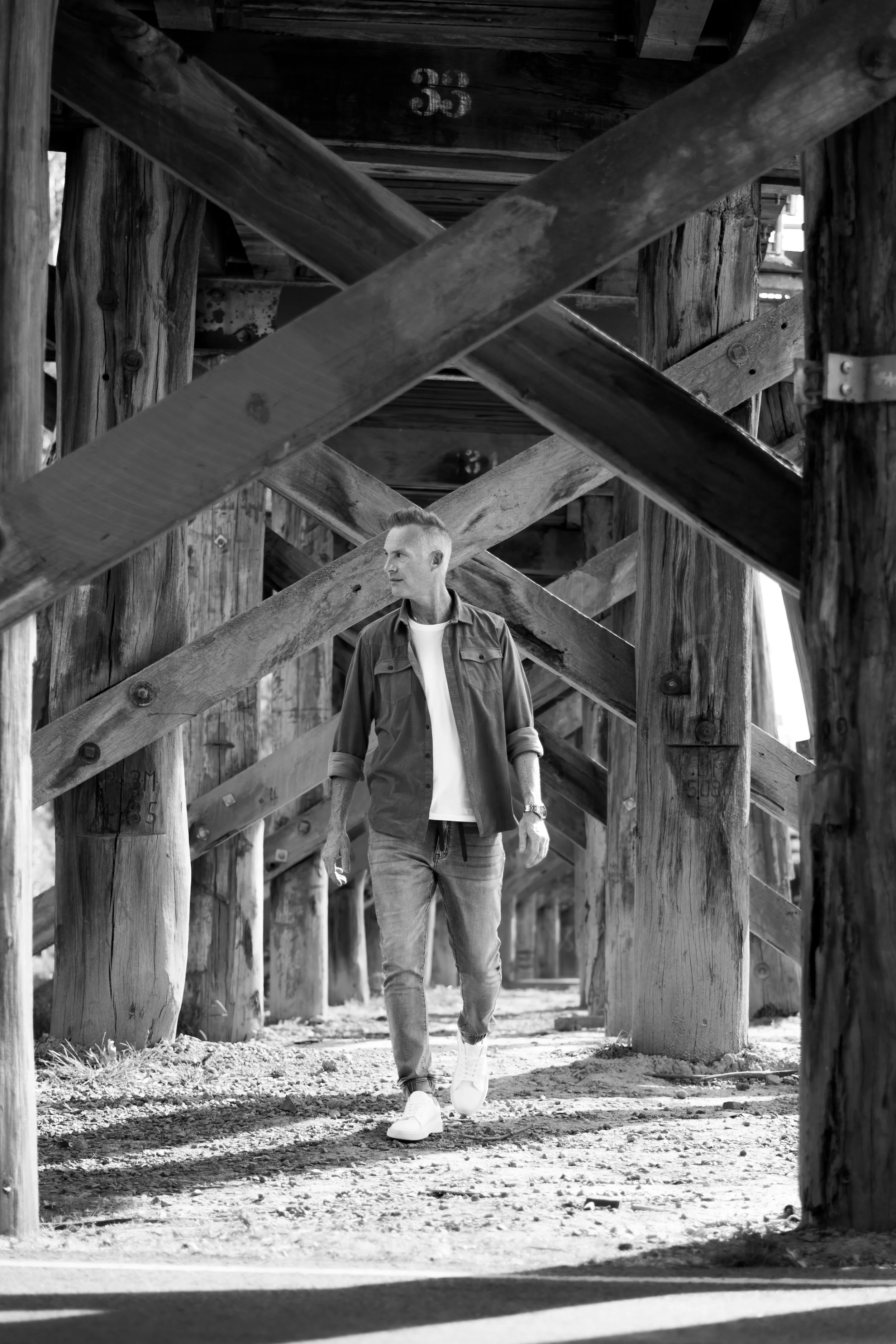 A man walking underneath a wooden bridge or pier structure, with wooden beams crisscrossing above and around him.