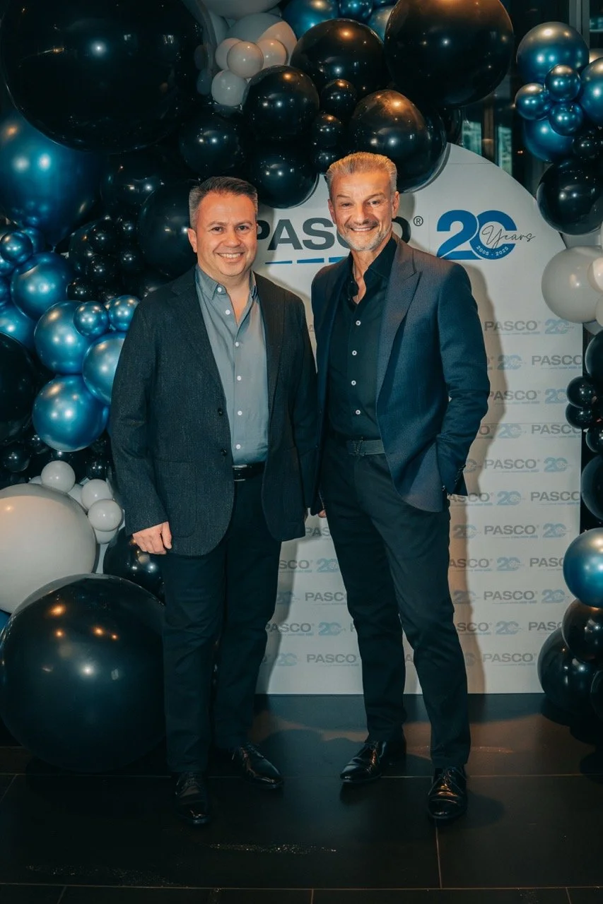 Two men in suits standing in front of a backdrop with PASCO and 20 years logo, surrounded by black, white, and blue balloons, at a celebration event.