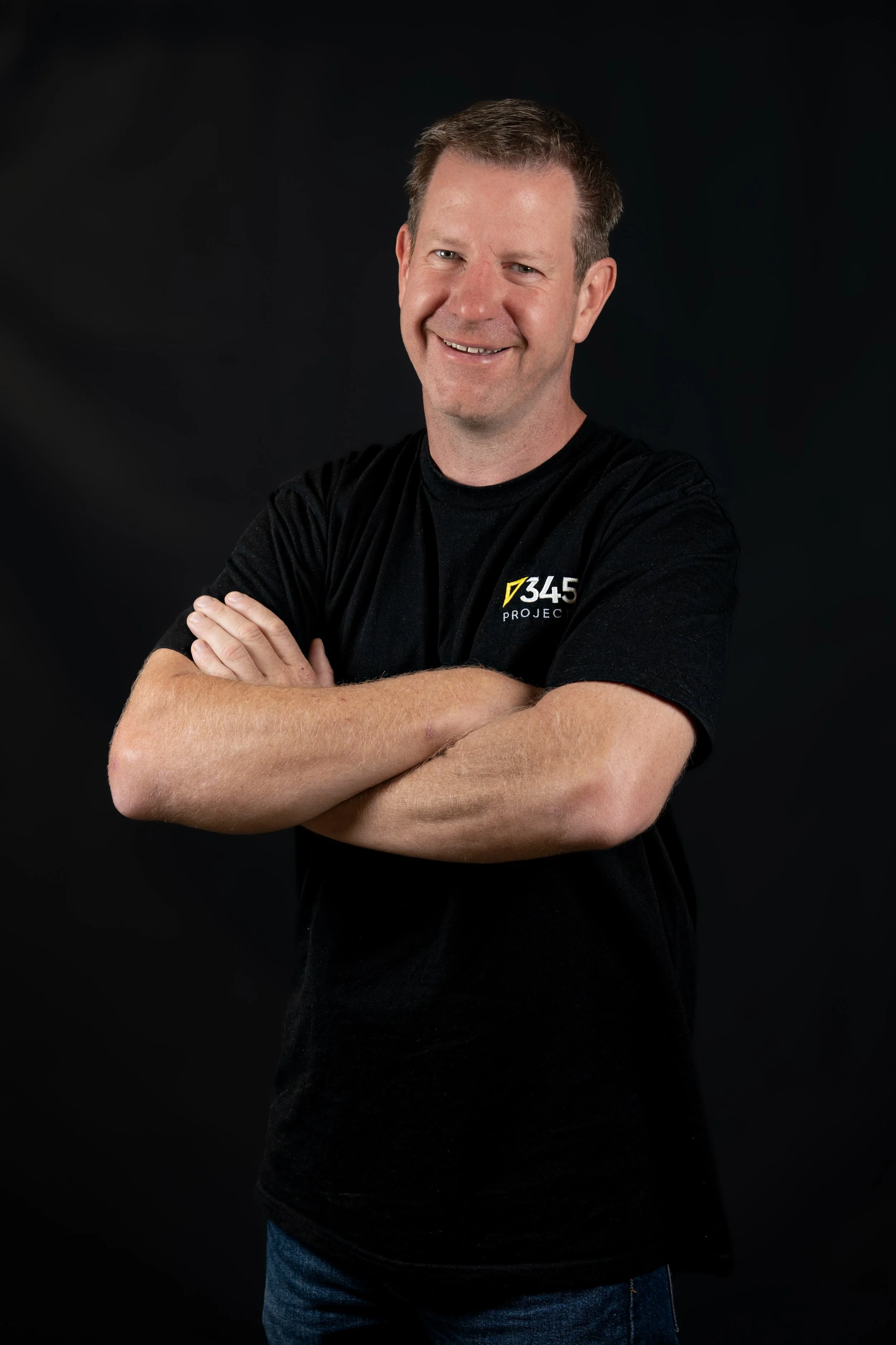A smiling man with short brown hair and fair skin, wearing a black T-shirt with '345 PROJECT' logo, arms crossed, against a dark background.
