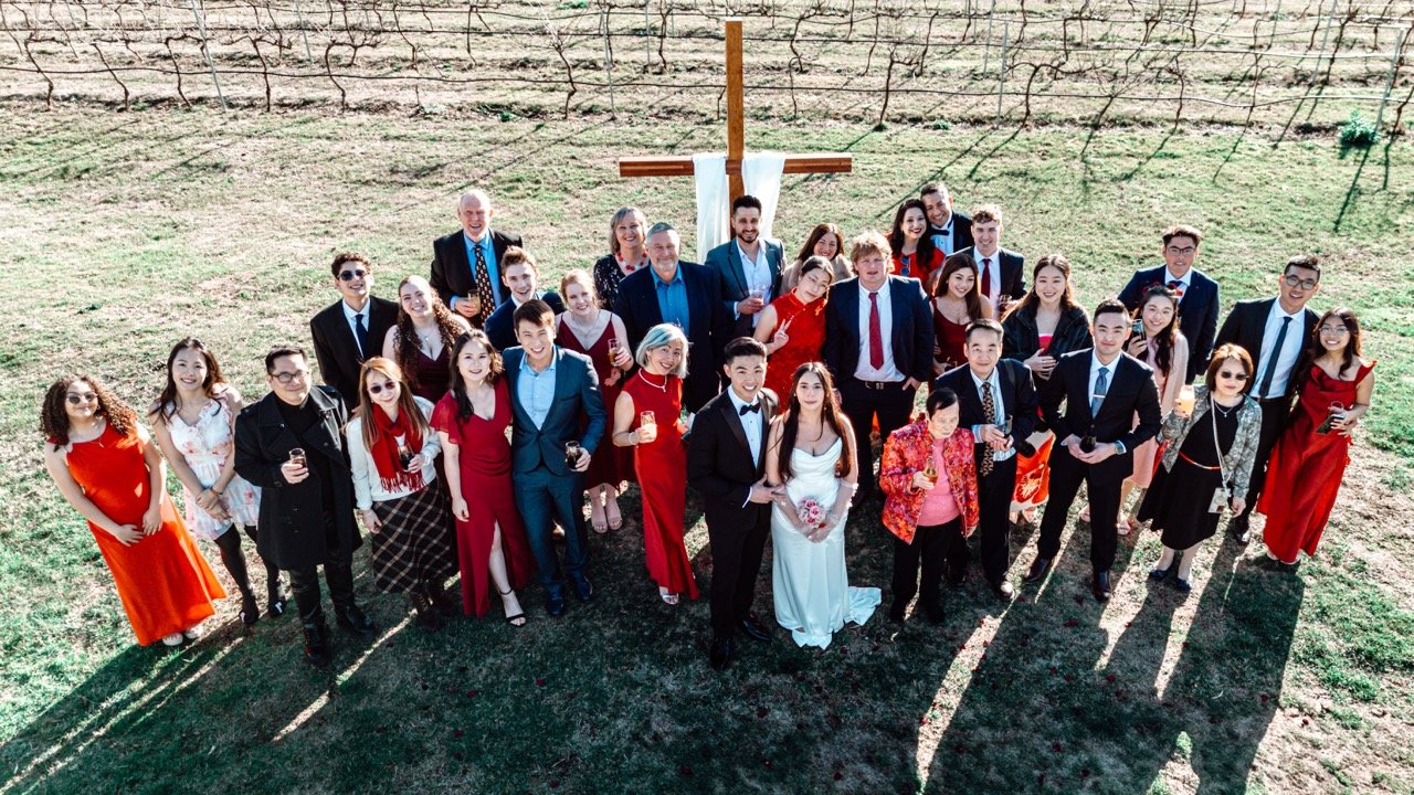 Group of people gathered outdoors for a celebration in front of a large wooden cross on a grassy hill, dressed in formal attire, some holding drinks, with sunny weather and clear skies.