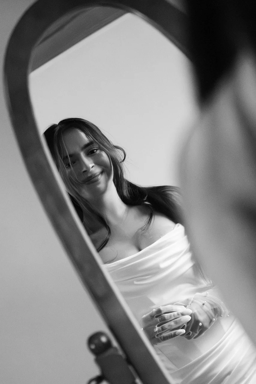 A woman is smiling while looking at herself in a mirror, taking a selfie. She is wearing an off-the-shoulder dress and has long dark hair.