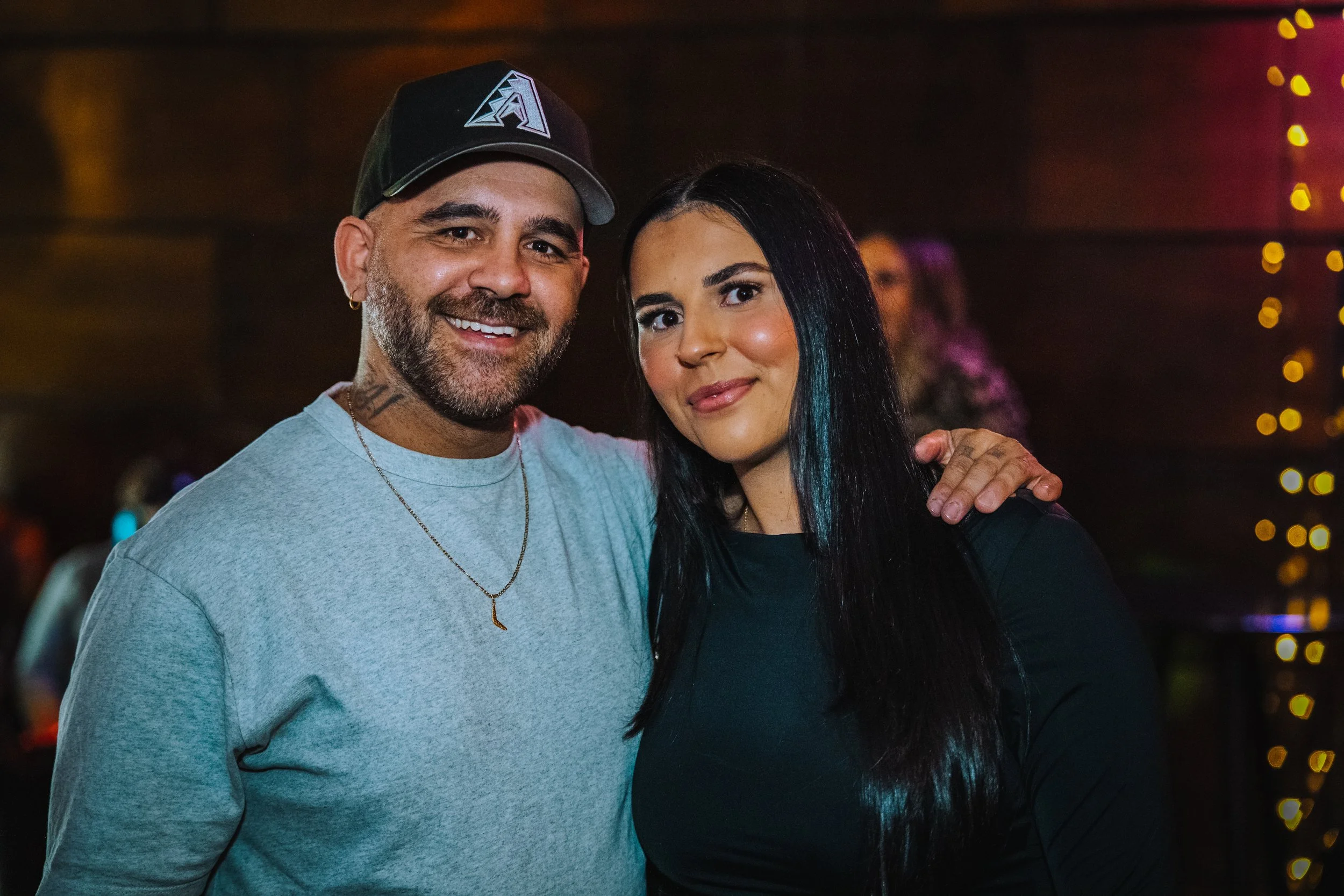A man and woman smiling at the camera at a social event with a dark background and bokeh lights.