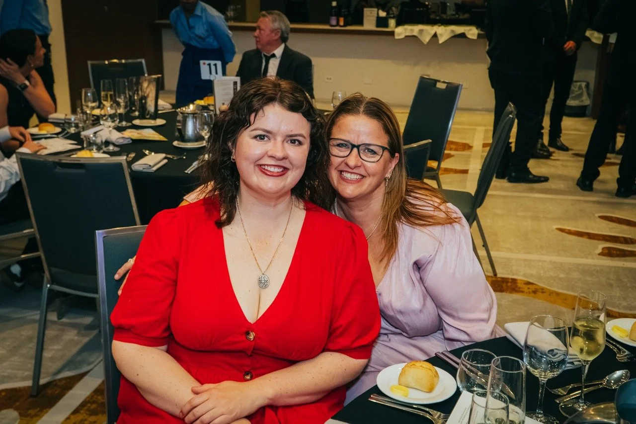 Two women smiling and sitting at a table during a formal event, with a banquet setting in the background.