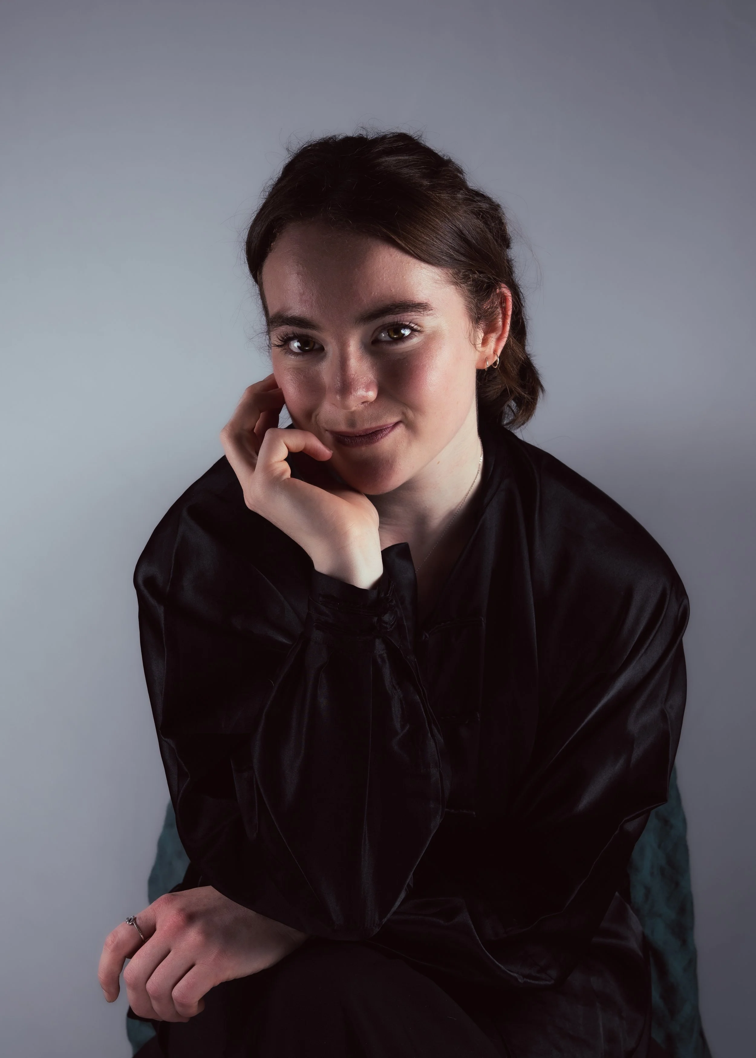 Portrait of a woman with dark hair, wearing a black satin jacket, sitting against a neutral gray background, smiling softly with her right hand touching her face.