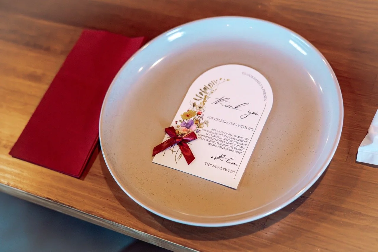 Empty white bowl on a wooden table with a red napkin beside it, containing a thank you note with floral design and a red ribbon.