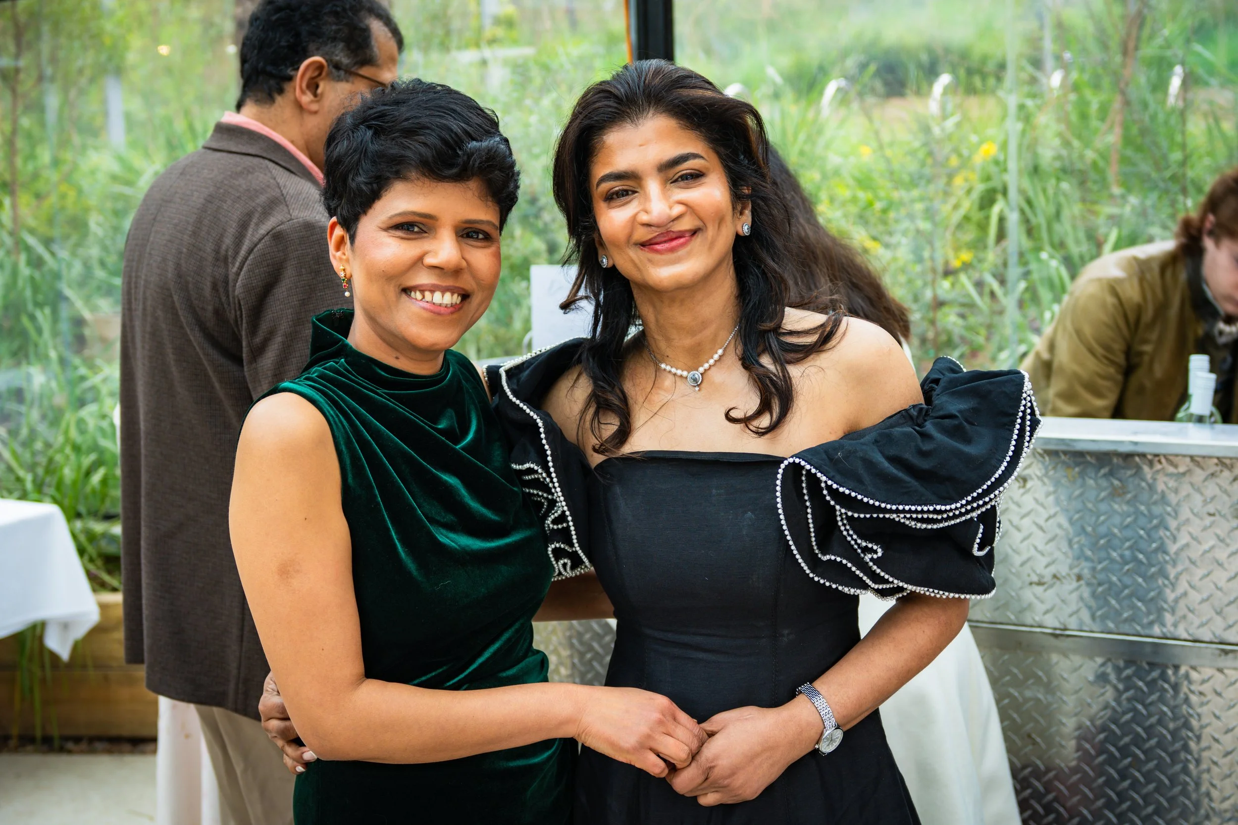 Two women smiling and holding hands at an outdoor event. One is wearing a dark green velvet dress, and the other is wearing a black dress with ruffled sleeves.