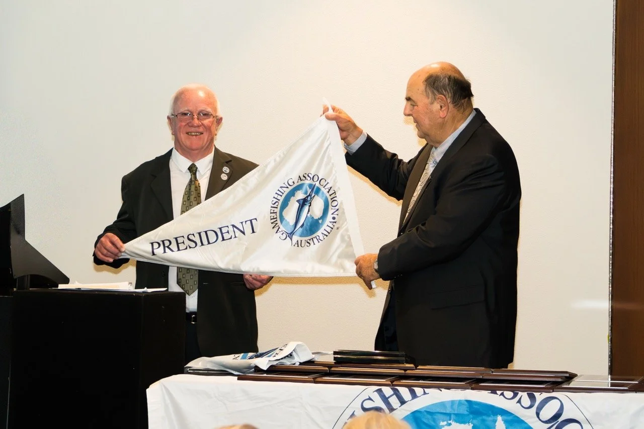Two men in suits, one holding a flag with the word 'PRESIDENT' and a logo, during a formal recognition or award ceremony.