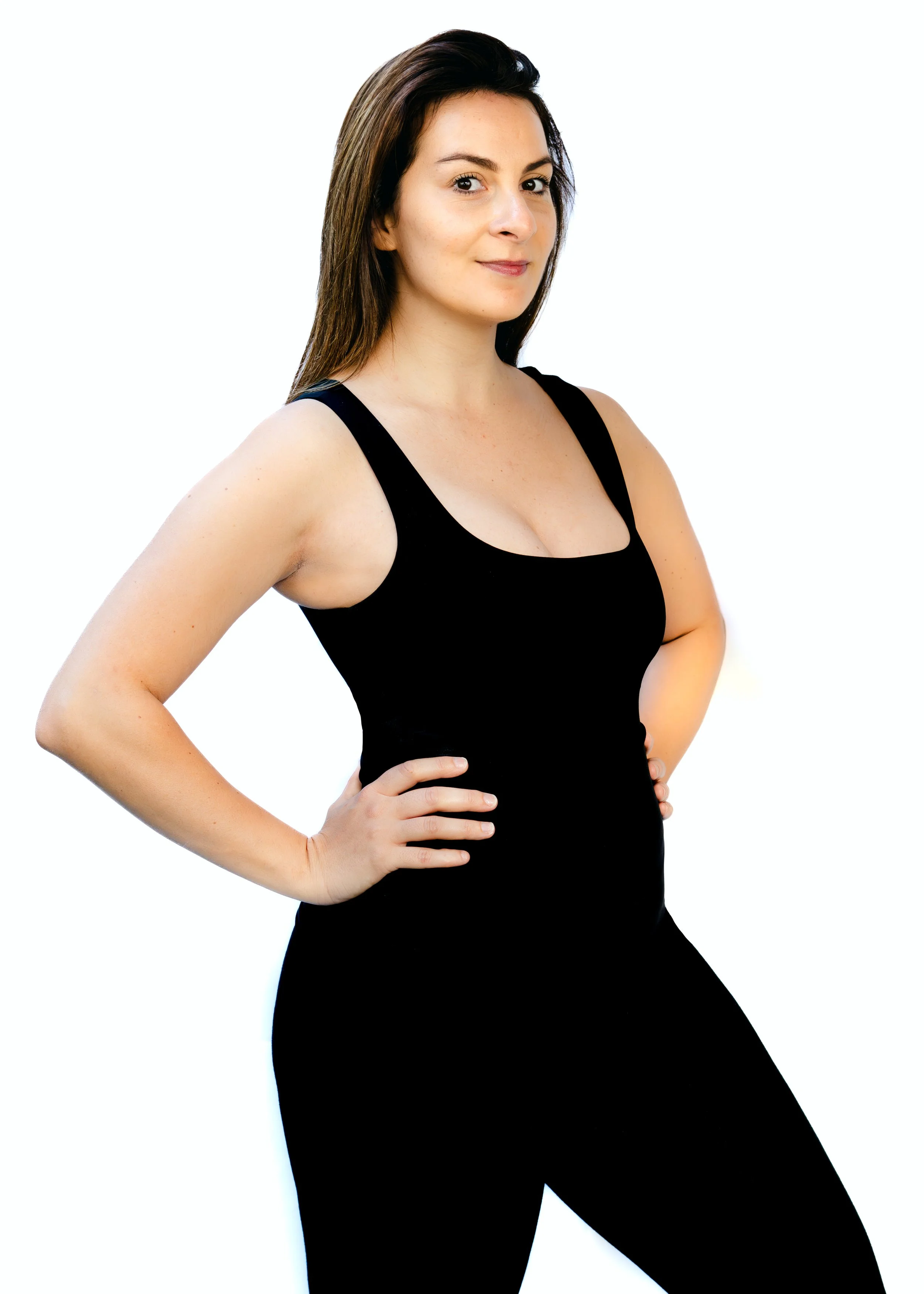 A woman with long brown hair wearing a black sleeveless top and black pants standing with her hands on her hips against a plain white background.