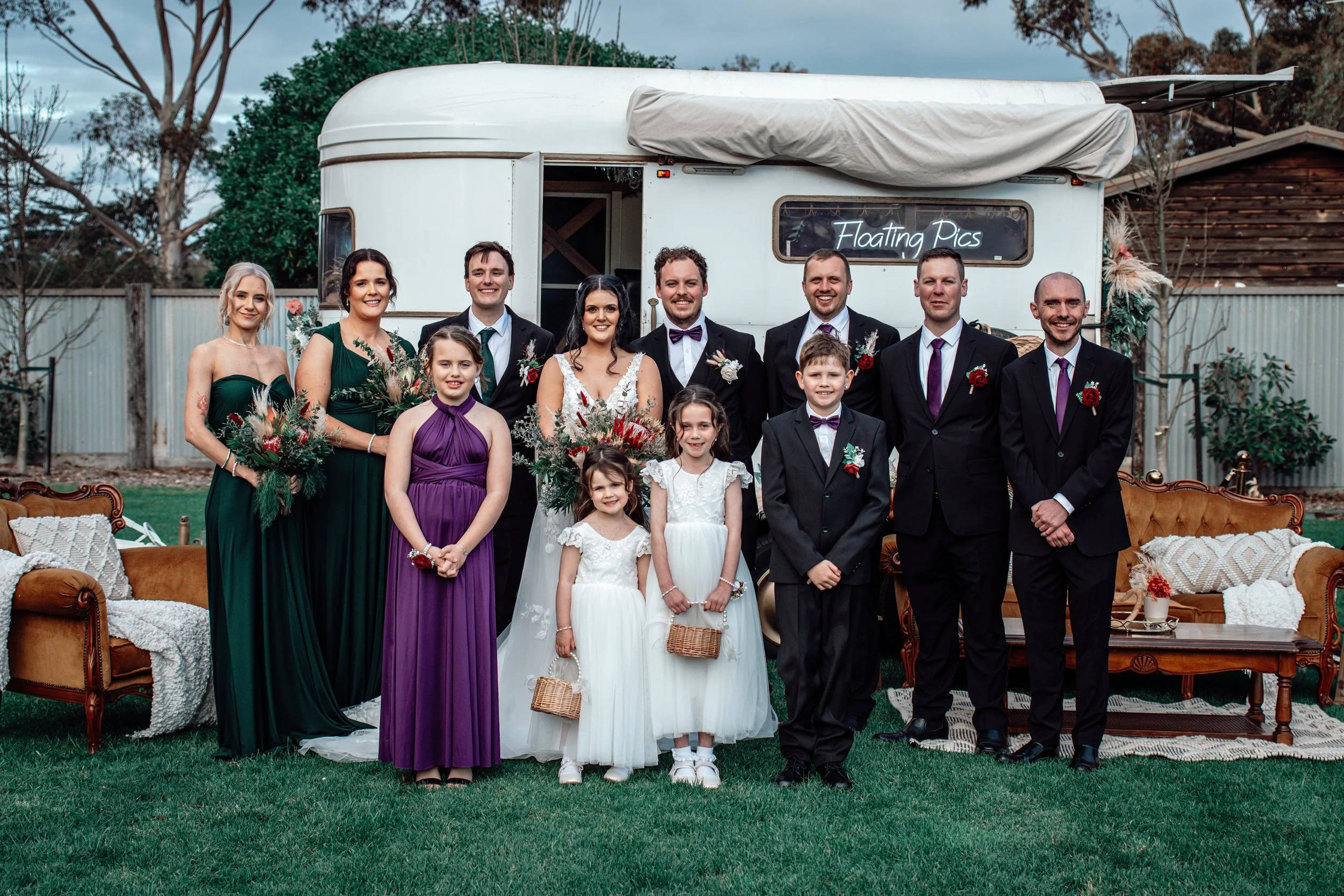A wedding party with bridesmaids, groomsmen, flower girls, and a bride and groom, posing outdoors in front of a vintage trailer with a sign that says ’Floating Pics’ during daytime.