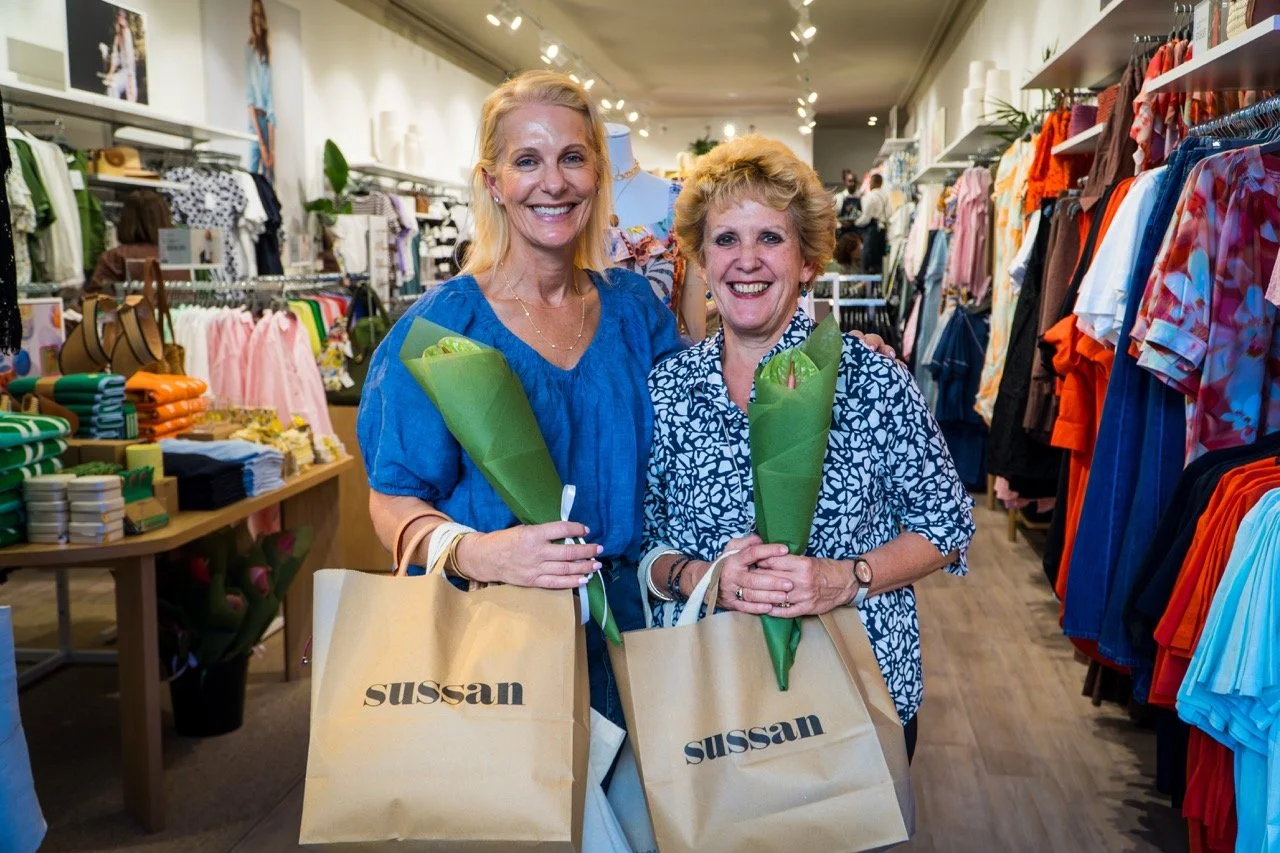 Two women smiling and holding bouquets of flowers in a clothing store, carrying beige shopping bags with the word 'Sussan' on them, surrounded by racks of colorful clothes.