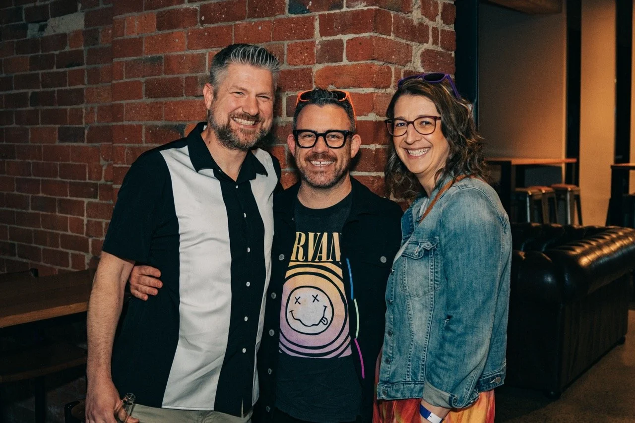 Three smiling adults standing close together in front of a brick wall in a dimly lit indoor setting.