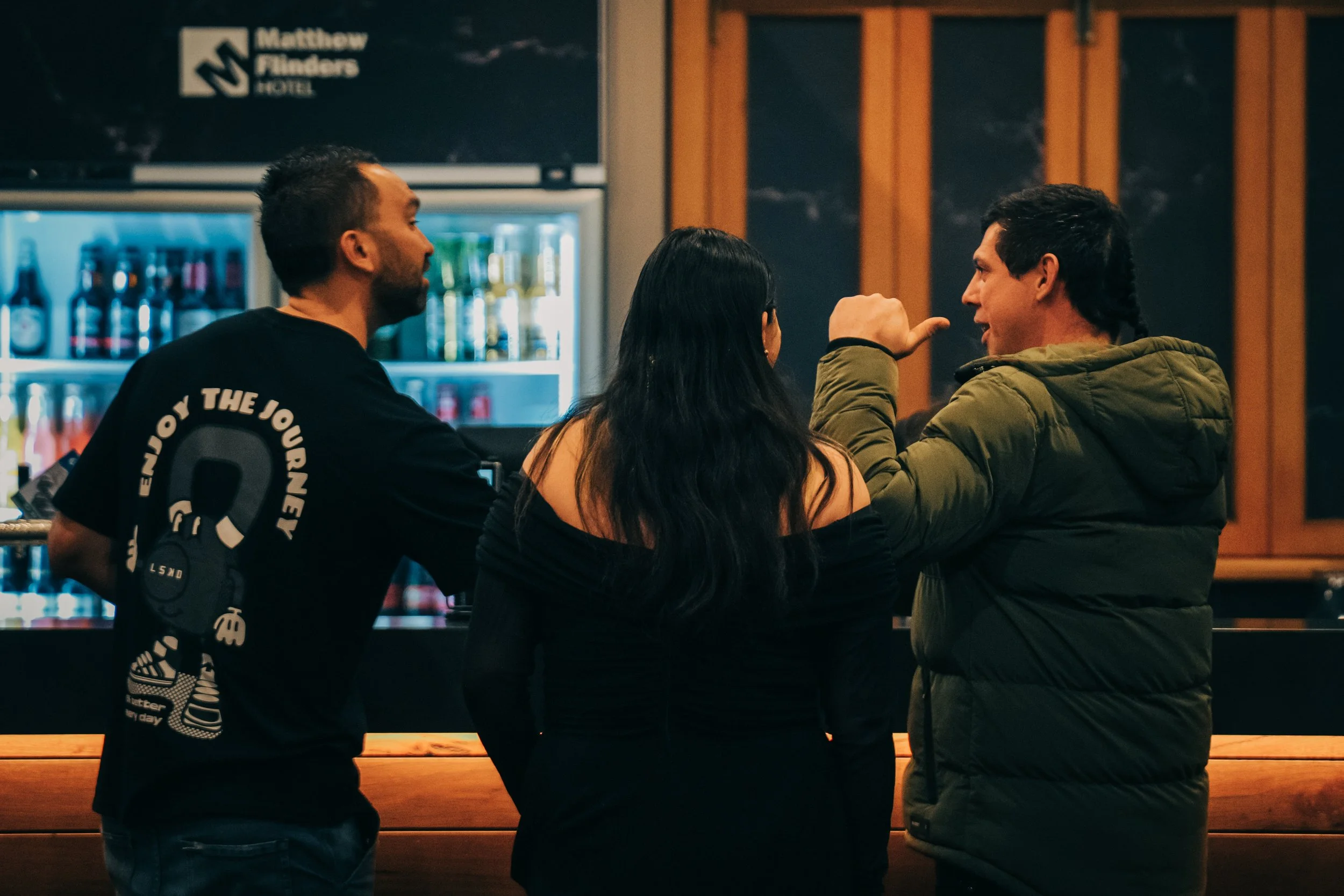 Three people are having a conversation at a bar; one man is pointing his finger and talking, the woman in the middle has long dark hair and is facing the man, and the man on the left is wearing a black t-shirt with white text, while the man on the ri