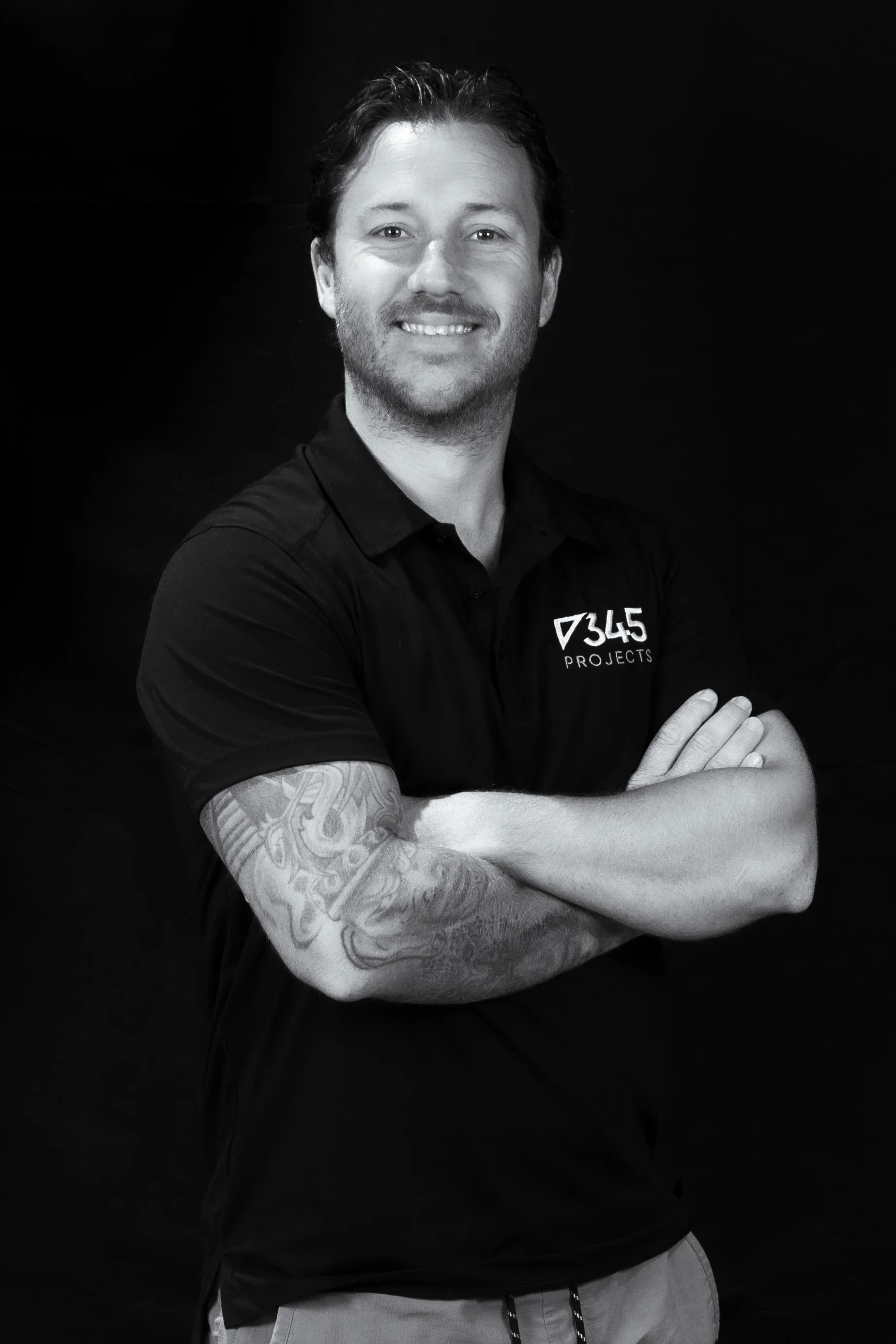 A man with tattoos on his left arm, smiling with arms crossed, wearing a black shirt with a logo that reads '345 PROJECTS', black background.