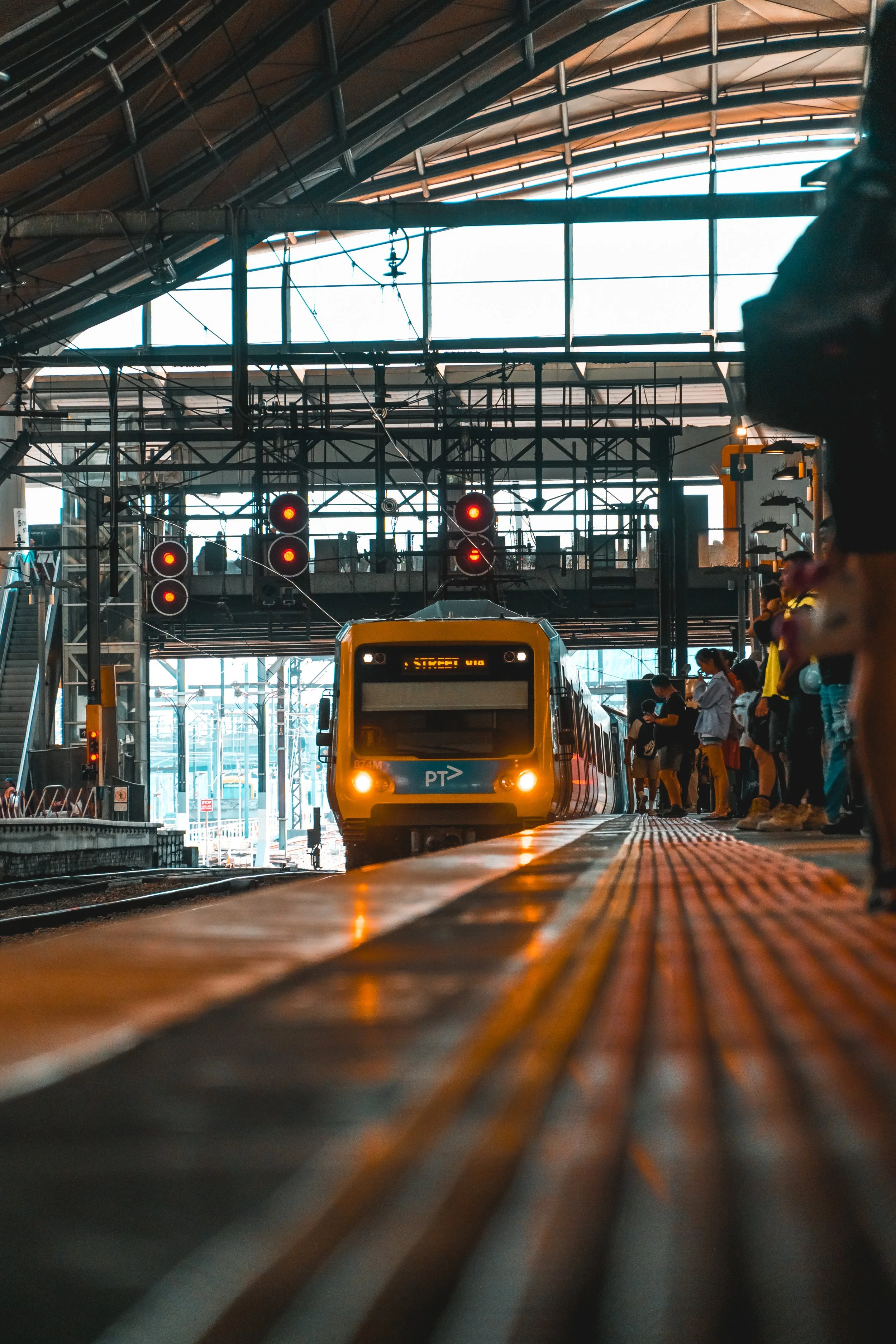 A yellow and blue train arriving at an indoor train station platform with passengers waiting, red signals overhead, and structural metal and glass roof.