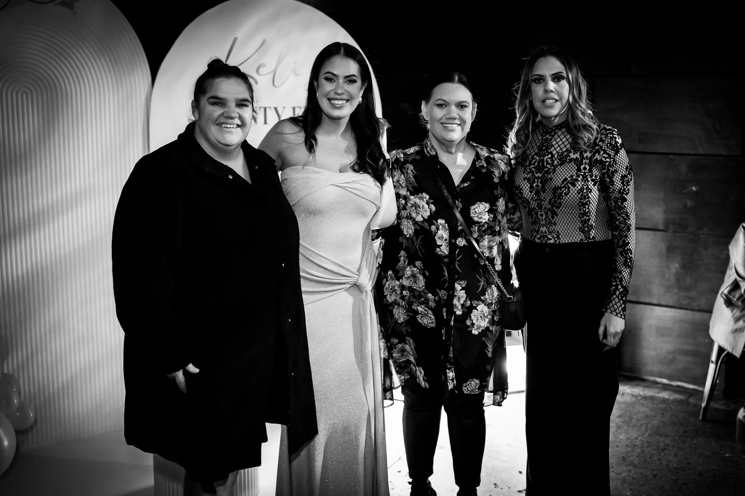 Four women standing together at an event, smiling for the camera, with a background displaying the words "Kelicia" and "Beauty."