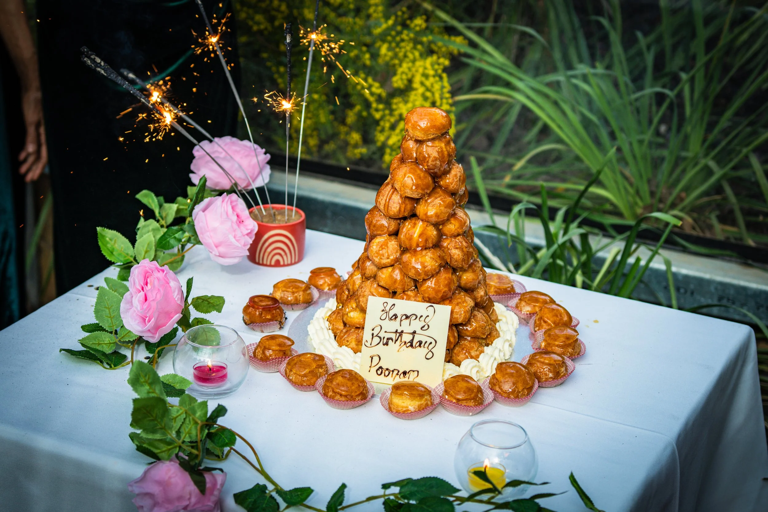A croquembouche birthday cake with pastries stacked in a cone and sparklers on top, decorated with pink roses and a sign that reads 'Happy Birthday Poornar,' on a table with candles and pink flowers.