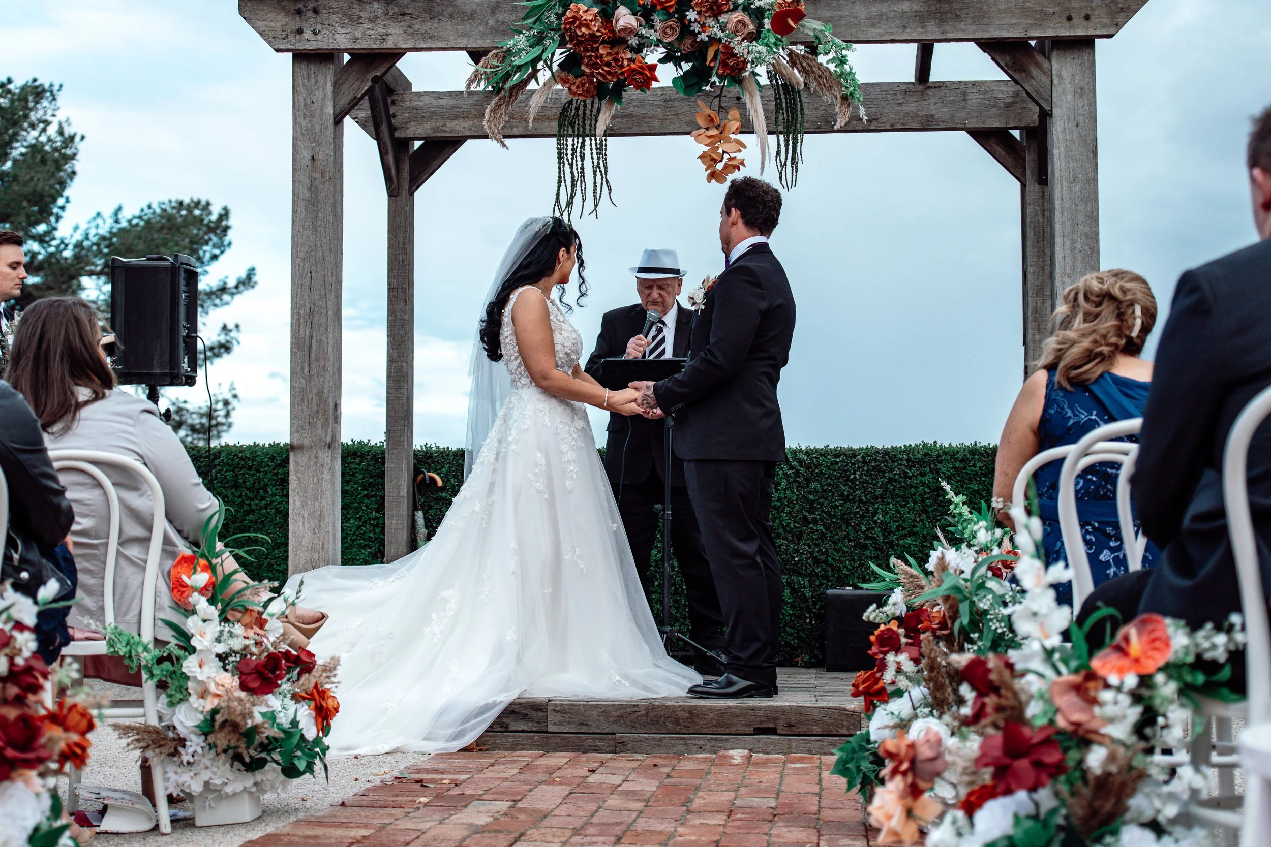 A couple getting married outdoors under a wooden arch decorated with flowers. The bride is in a white wedding gown with a veil, and the groom is in a black suit. An officiant, an older man with a hat, is reading from a book. Guests are seated on whit