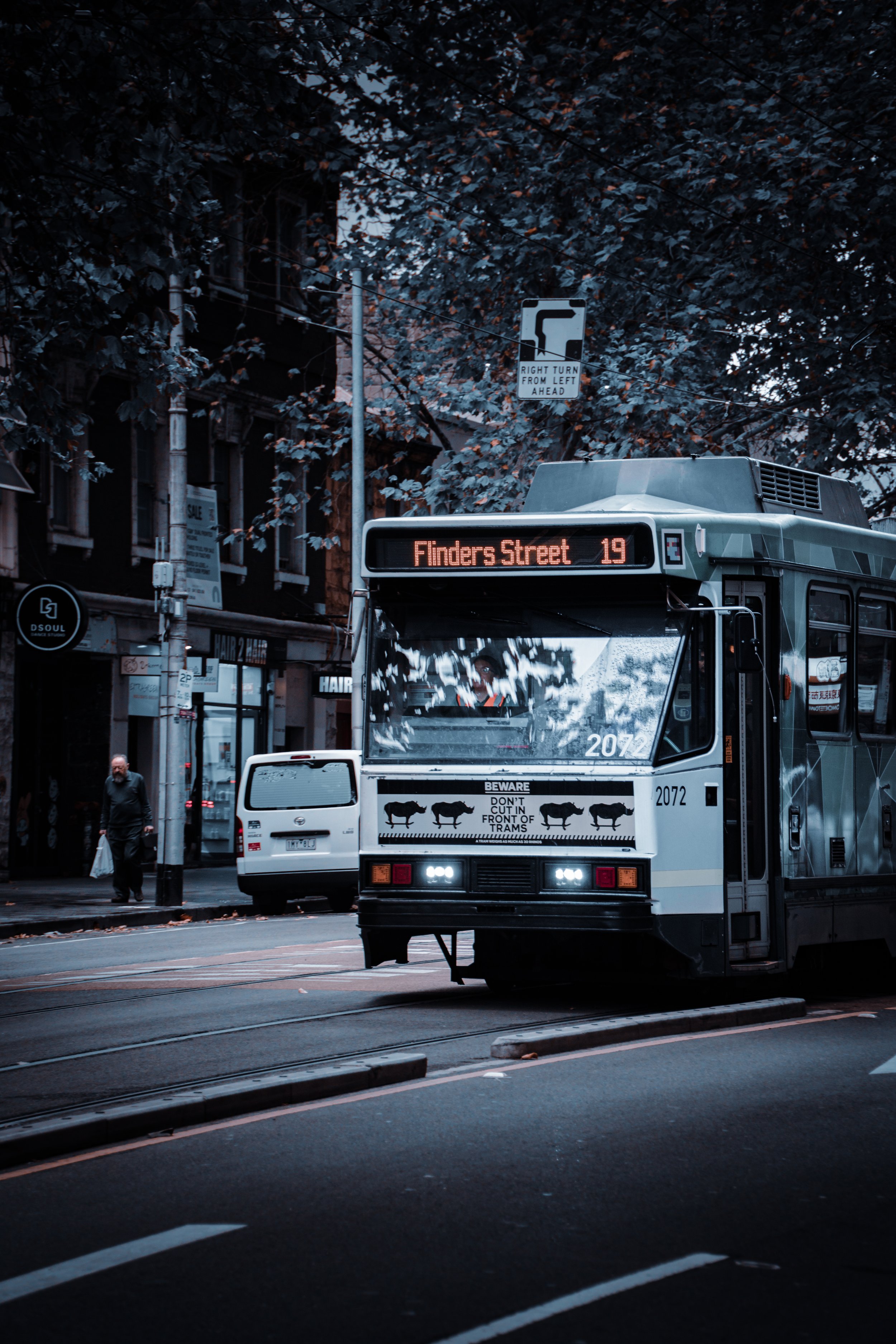 A city street scene featuring a tram with a sign reading 'Finders Street 19' and a URL warning about donkeys in front of tram tracks. Pedestrians walk along the sidewalk, with a white vehicle parked nearby and trees overhead.