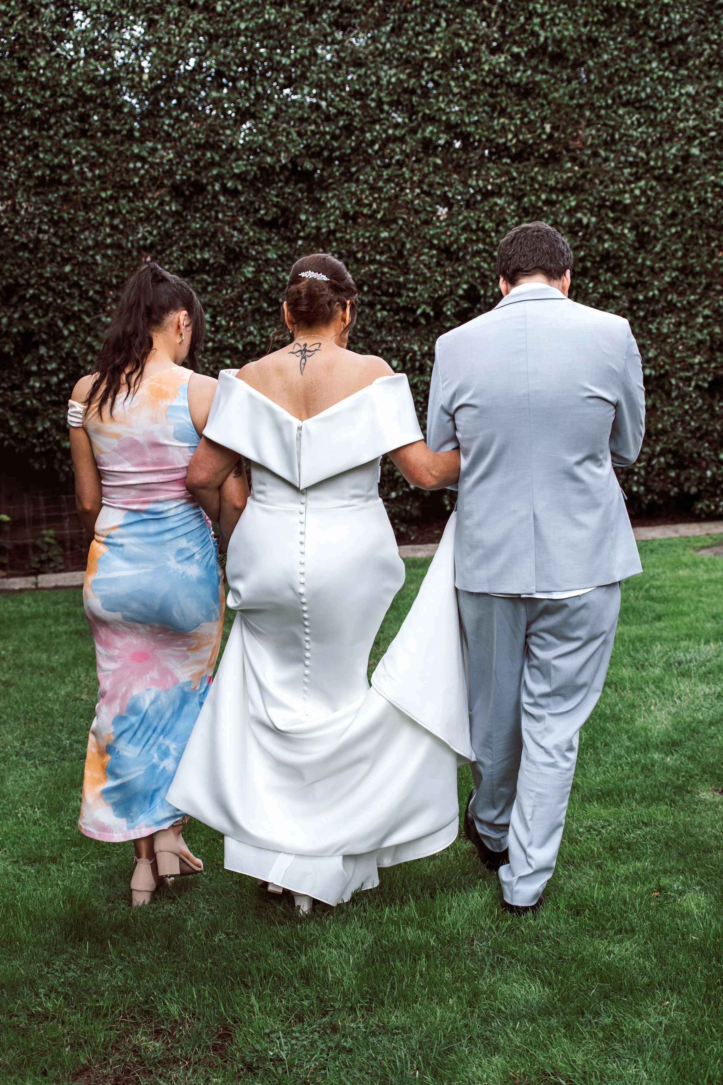 Back view of a bride in a white wedding gown walking with a groom in a light gray suit, and a woman in a colorful dress, all holding each other's arms in a garden with green bushes.