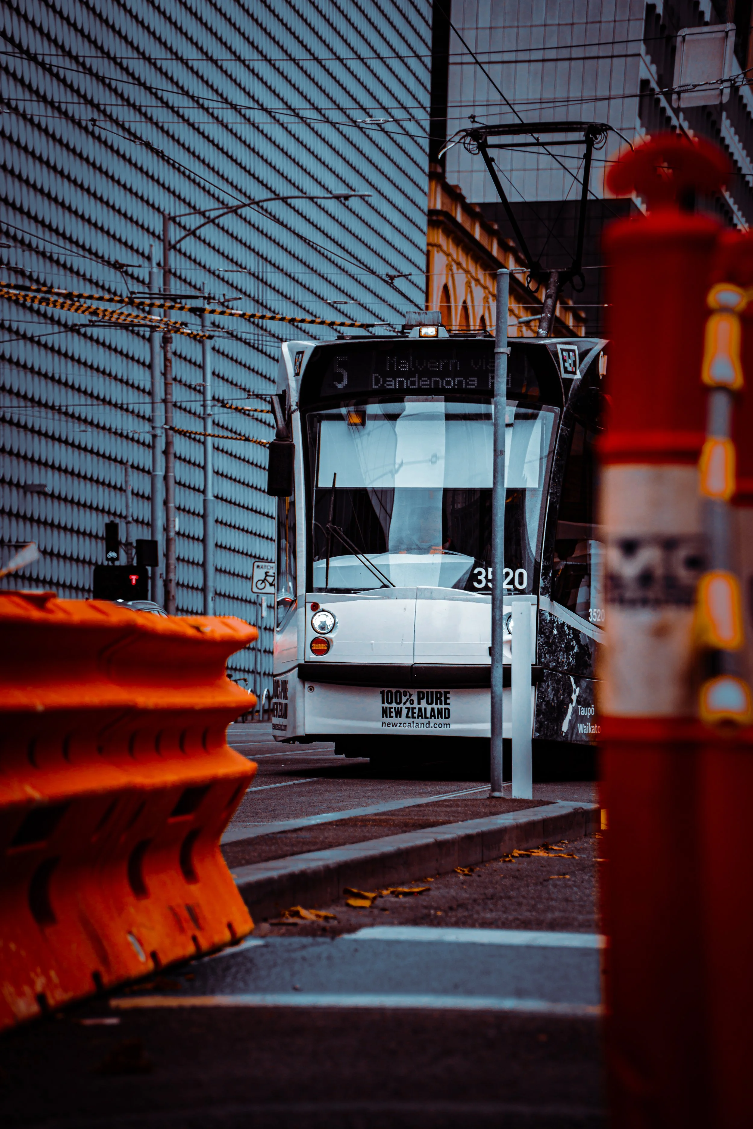 A modern tram traveling on a city street, with orange safety barriers and street construction signs in the foreground.