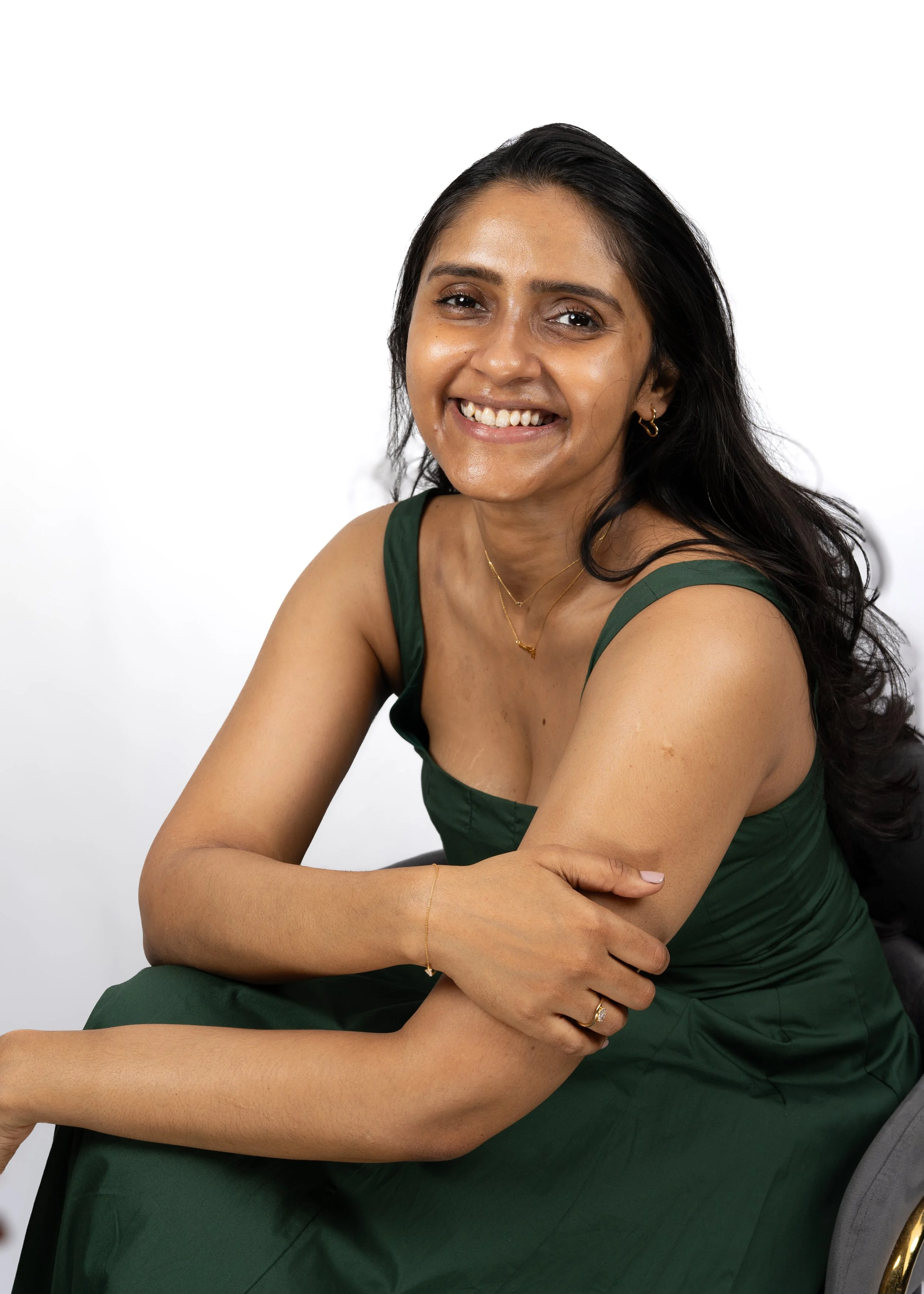 Smiling woman with dark hair and a green dress sitting with arms crossed, against a white background.