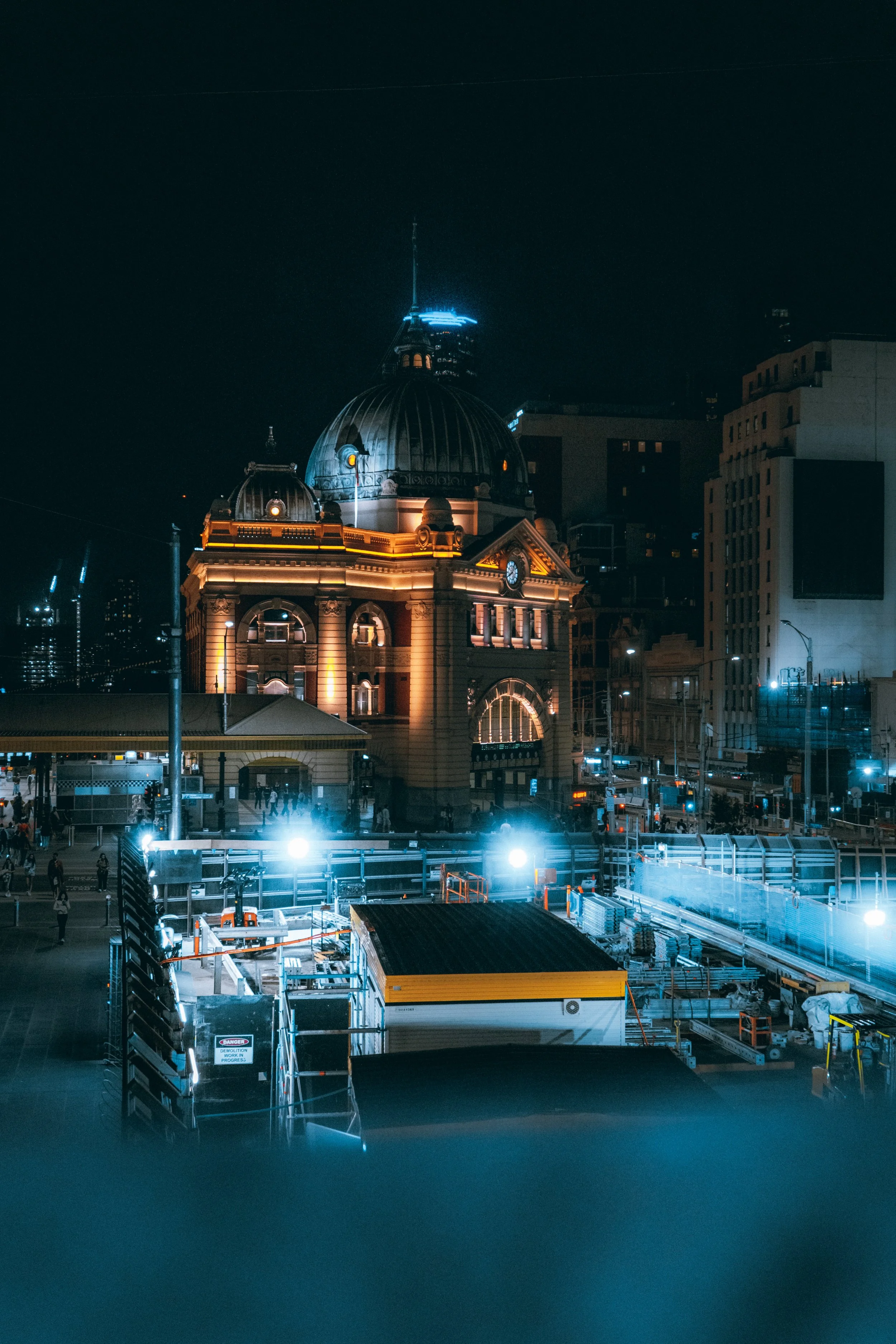 Nighttime view of a historic building with a domed roof, illuminated with orange and white lights, in a cityscape with construction site and tall buildings in the background.