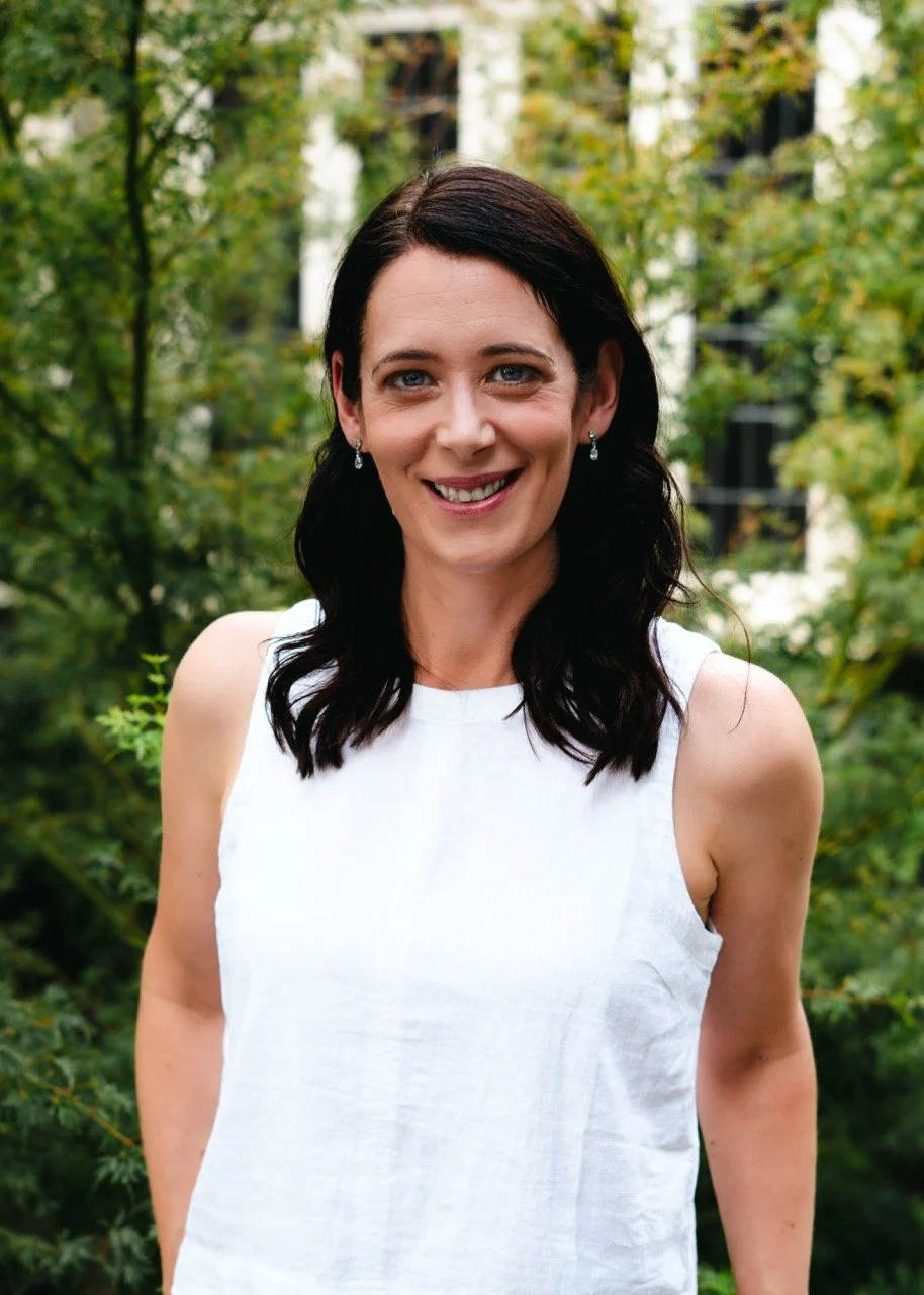 A woman with shoulder-length dark hair and blue eyes, smiling, wearing a sleeveless white top and earrings, standing outdoors with green foliage in the background.