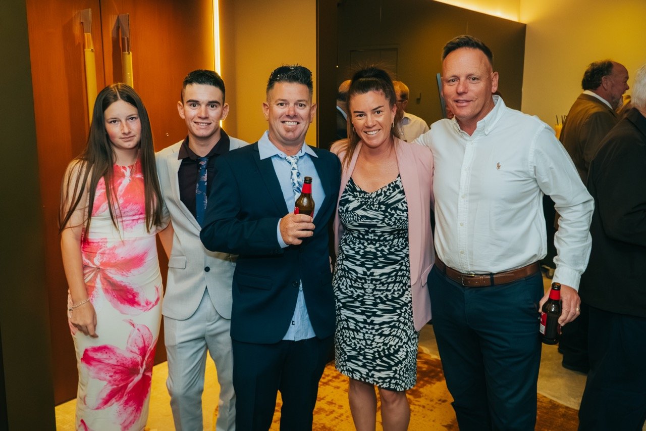 Group of five people standing together at a social event, smiling, with two holding beer bottles.
