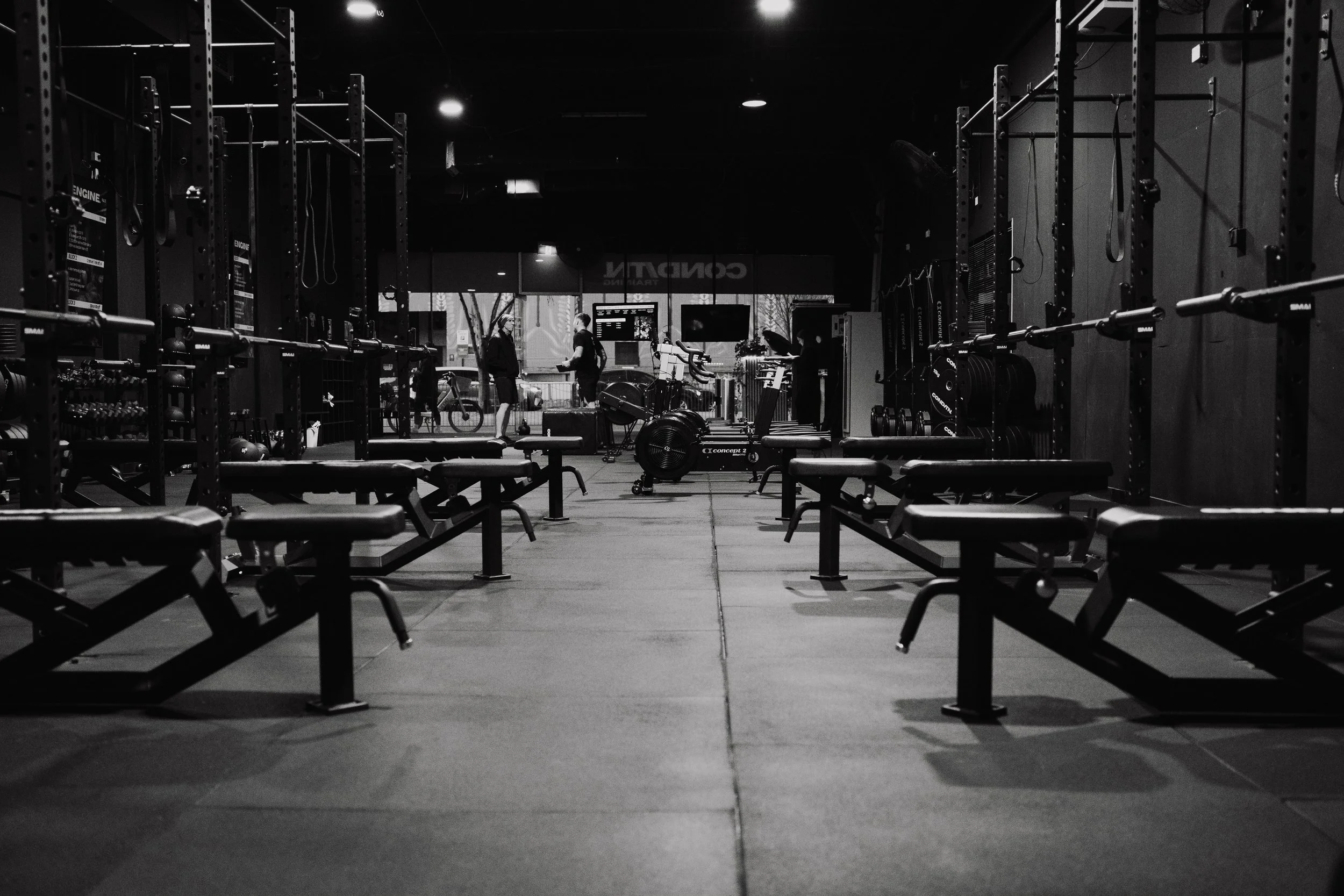 Empty gym with black workout benches, weight racks, and exercise equipment, viewed through a wide aisle, with three people talking in the background near large windows.