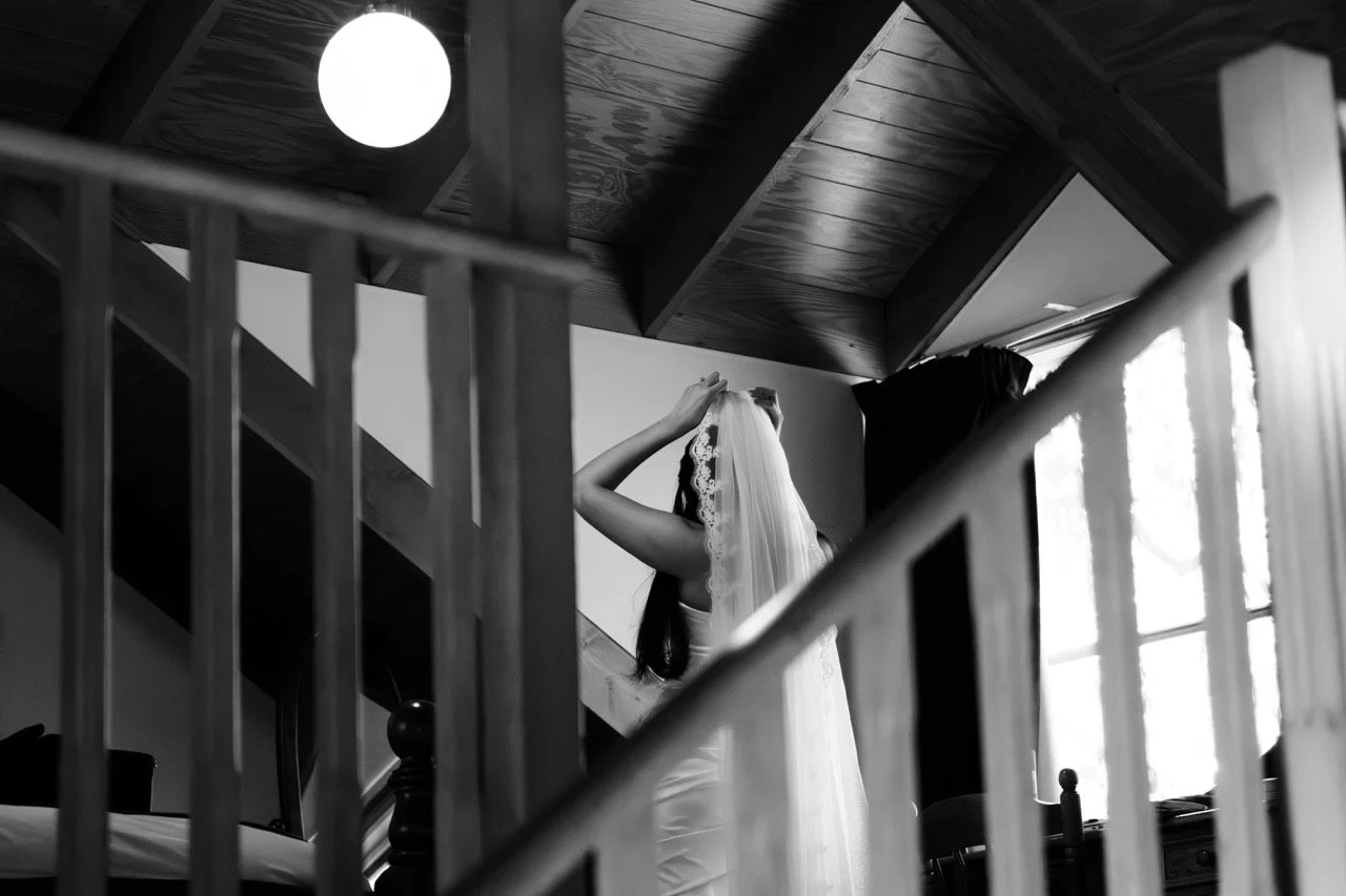 A bride in a wedding dress and veil standing on stairs, adjusting her veil, with their face not visible. The scene is in a room with wood-paneled ceiling, a window with curtains, and a ceiling light.