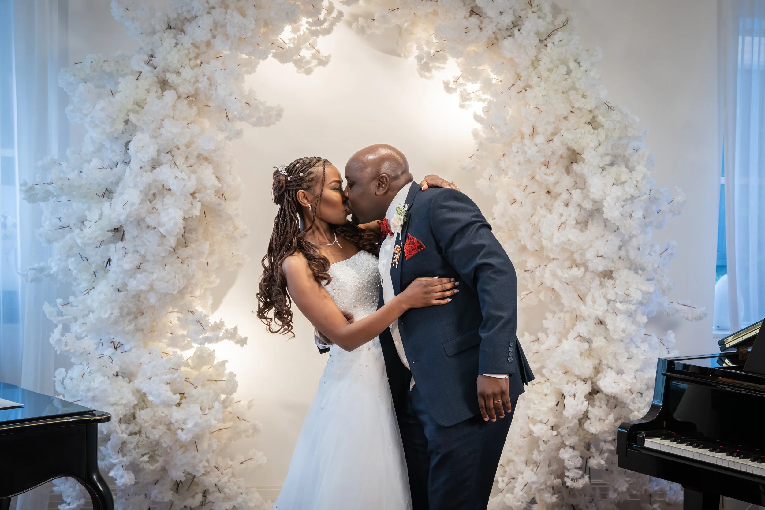 A bride and groom share a kiss during their wedding ceremony in front of a white floral arch. The bride has long, braided hair and is wearing a white wedding gown, while the groom is bald and dressed in a dark suit with a red pocket square and bouton
