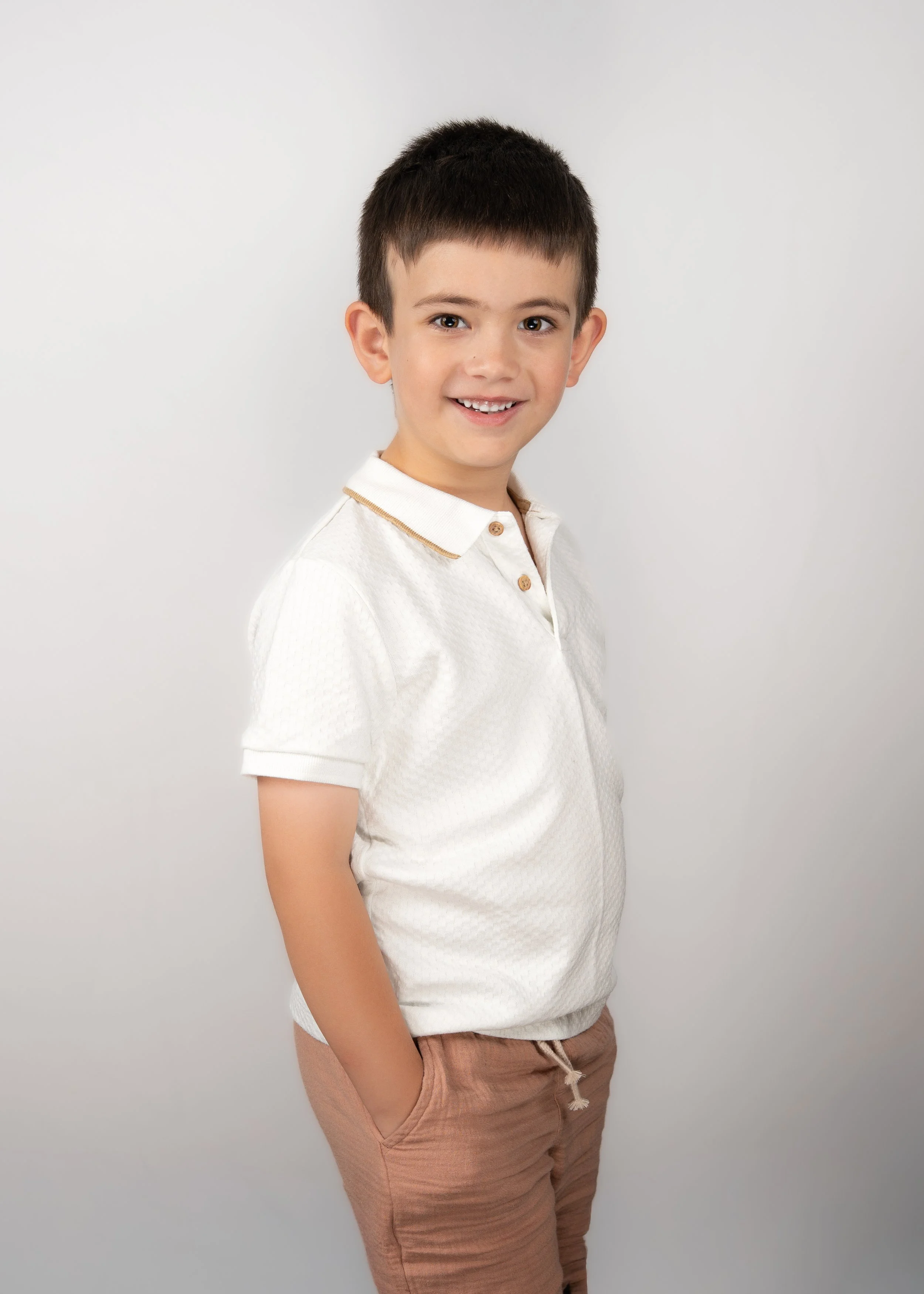 A young boy with short dark hair smiling, wearing a white polo shirt and tan pants, standing against a plain light gray background.