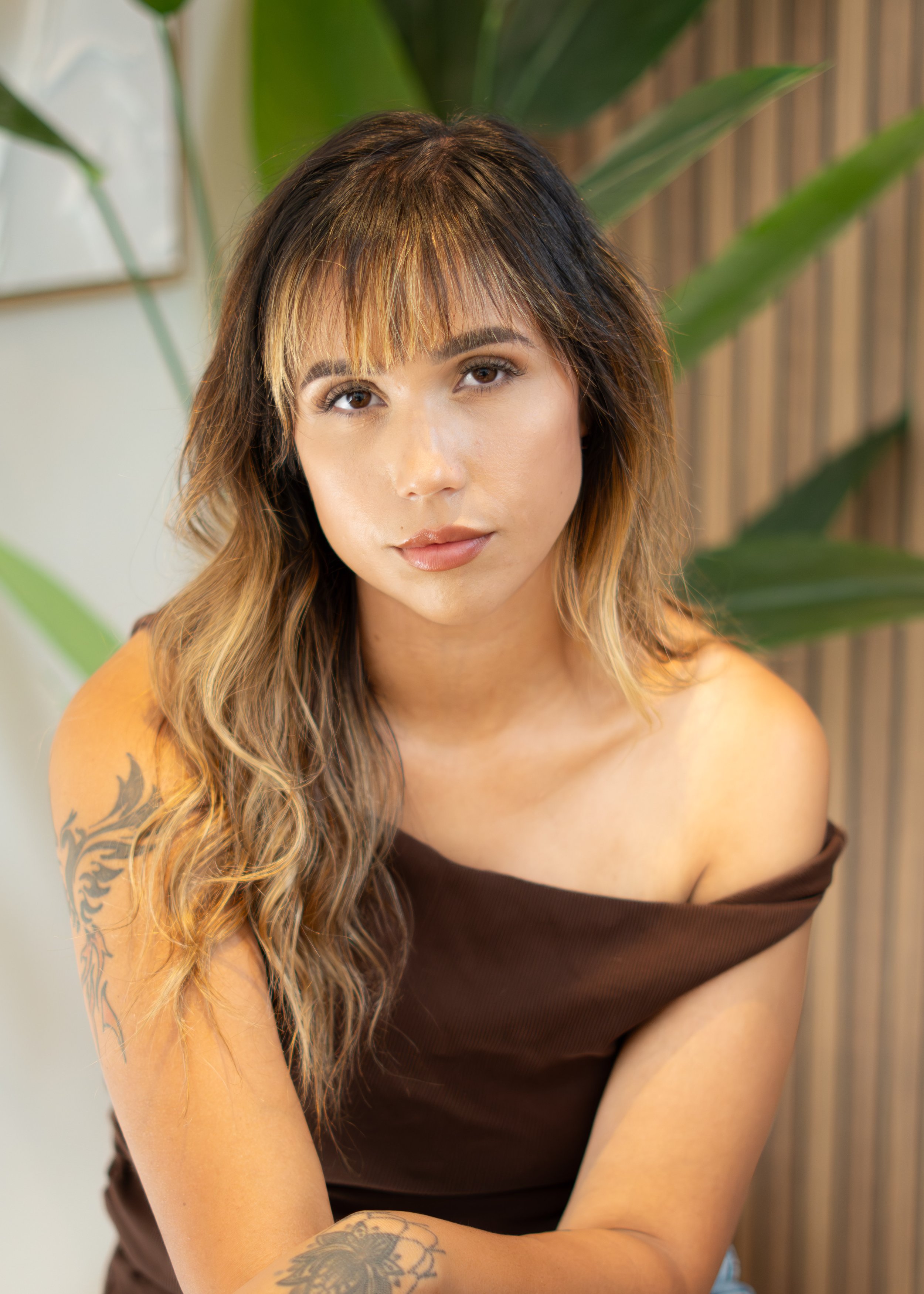 A young woman with long, wavy hair and tattoos on her arm, wearing a brown off-shoulder top, looks into the camera with a neutral expression. Green leaves and a wooden background are visible behind her.