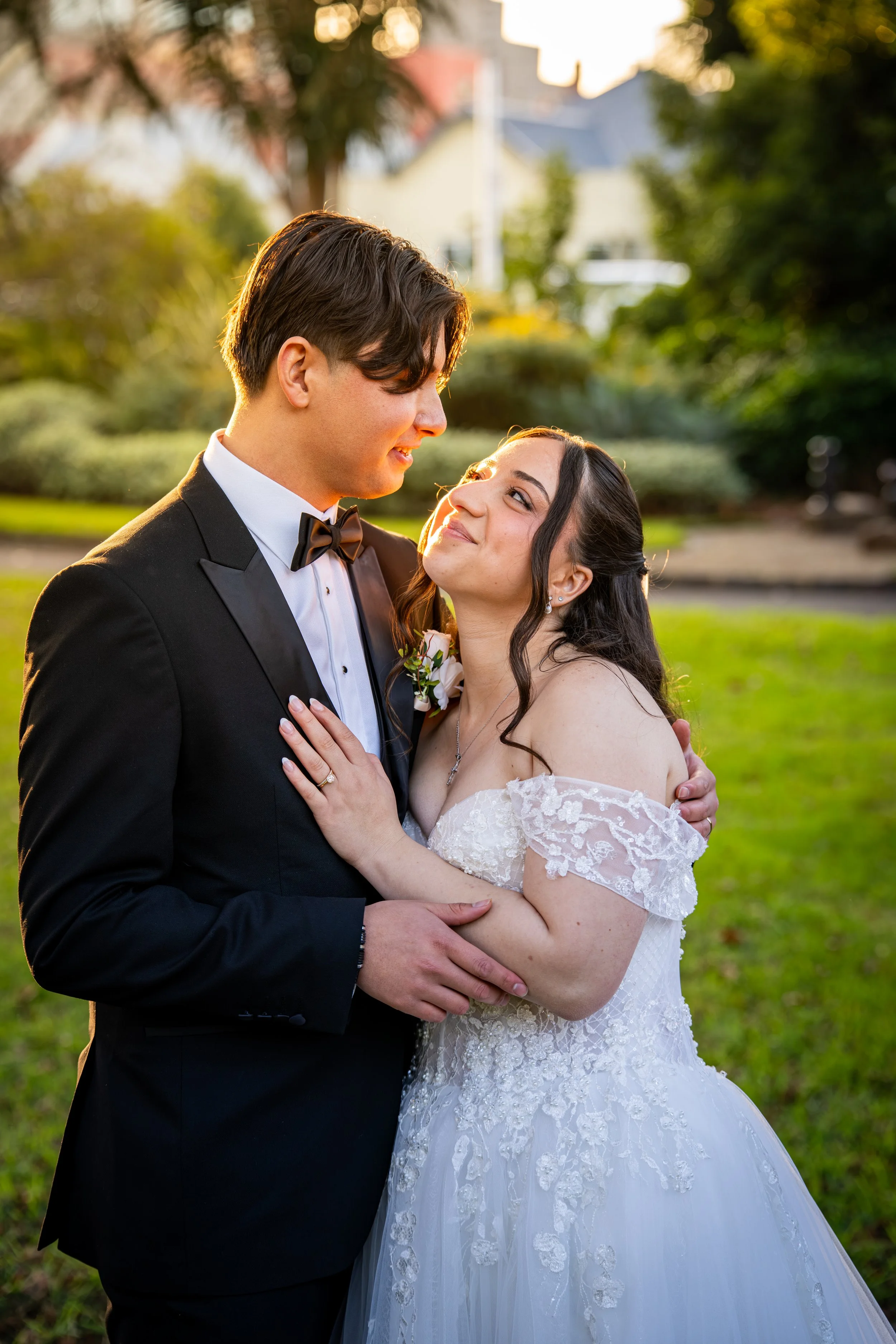 A newlywed couple stands close together outdoors, embracing at sunset. The groom wears a black tuxedo with a white shirt and black bow tie, and the bride wears a white lace wedding gown with off-the-shoulder sleeves. They are gazing into each other's
