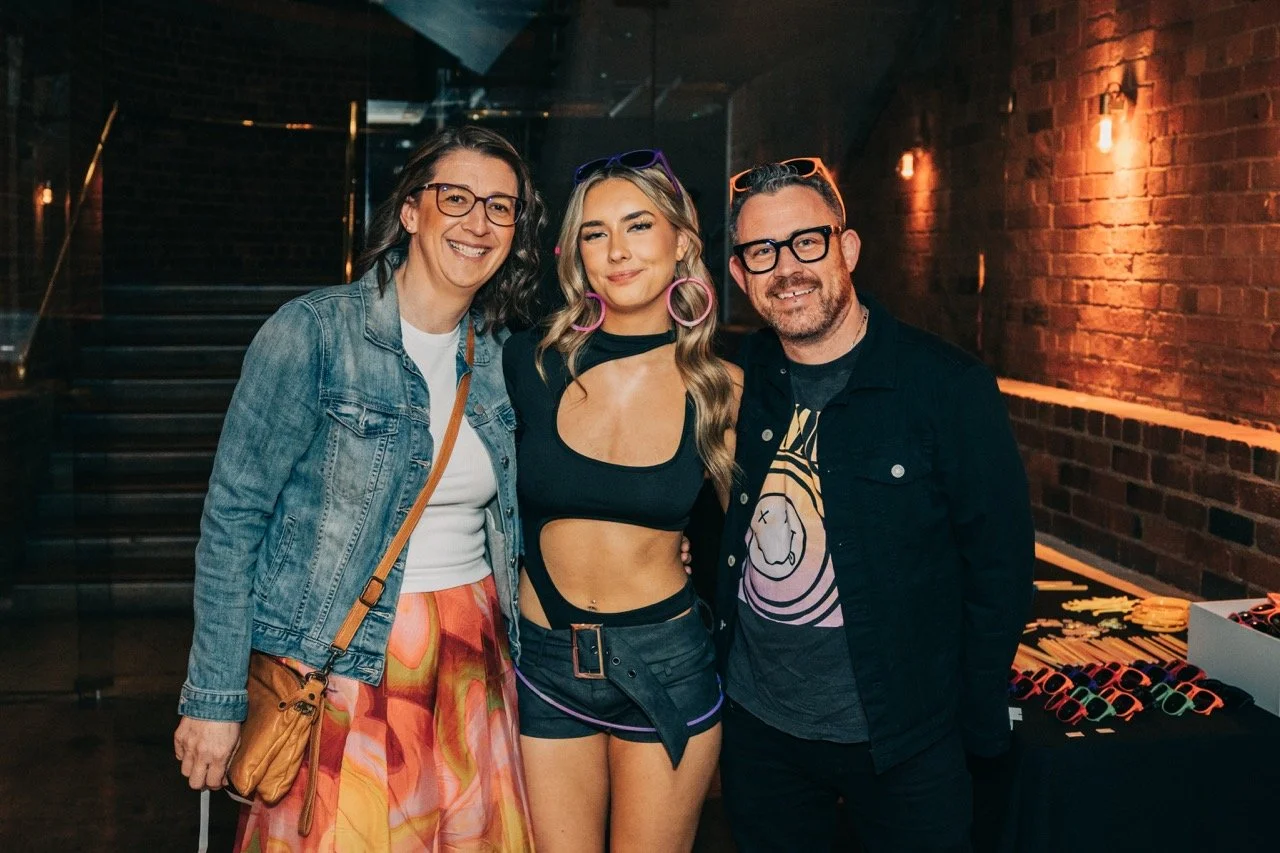 Three people, two women and one man, posing together indoors at a social event. The woman in the middle is wearing a black crop top, black shorts, and colorful accessories. The woman on the left is wearing glasses, a denim jacket, and colorful pants.