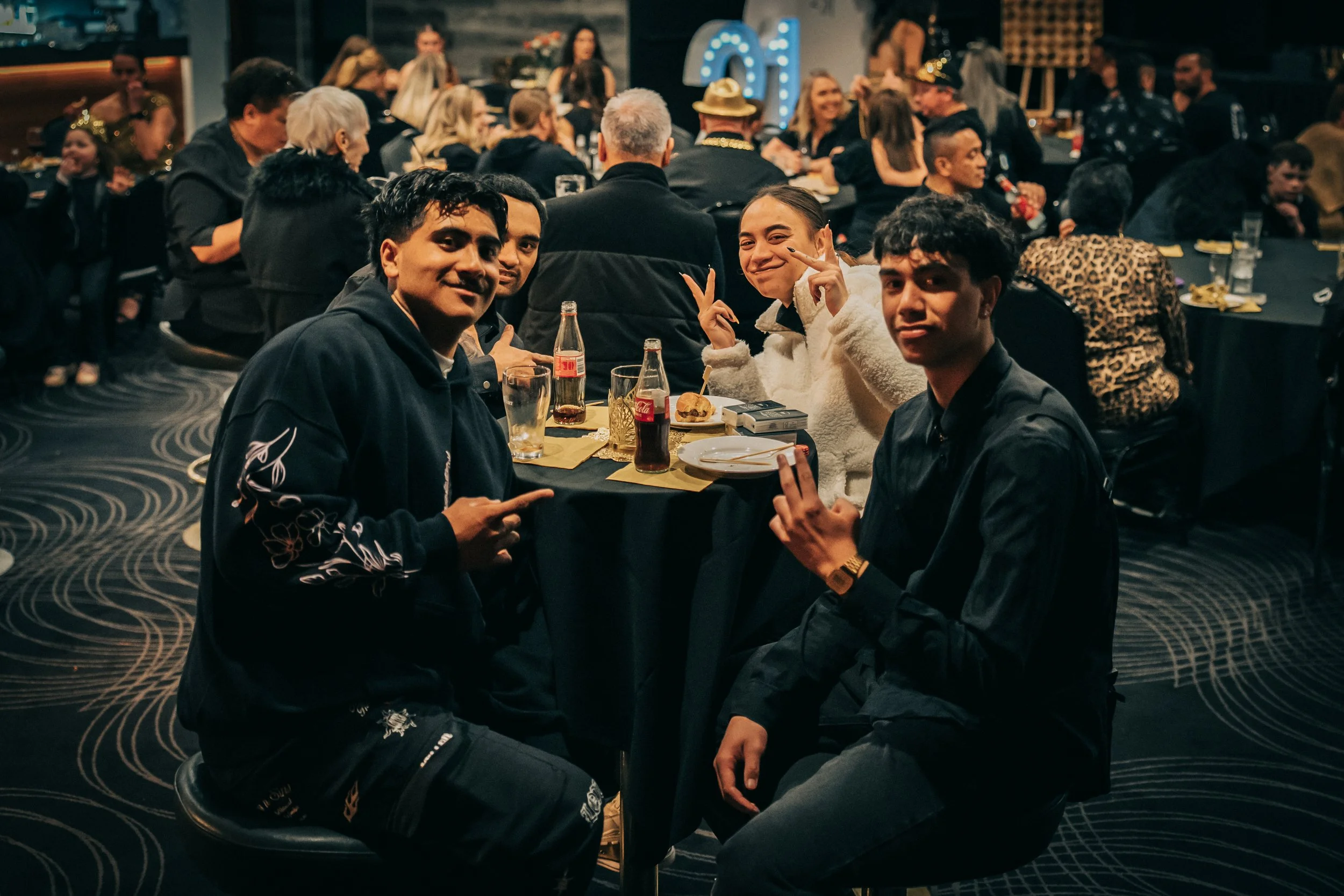 Group of four young people sitting at a table during a festive indoor event, with other guests in the background, some wearing party hats and casual attire, and a lit decorative number '01' in the background.