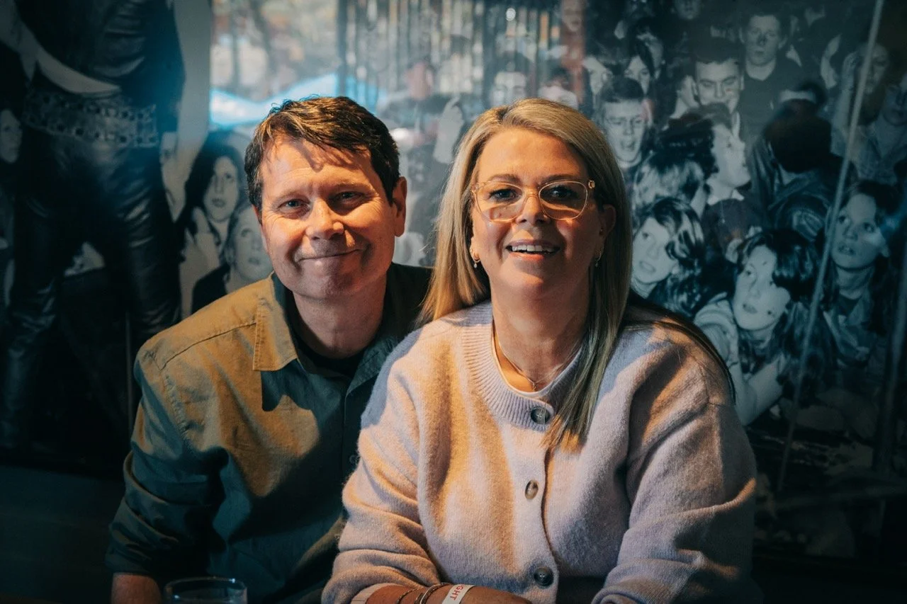 A man and woman sitting together in front of a black and white mural of numerous people, smiling at the camera.