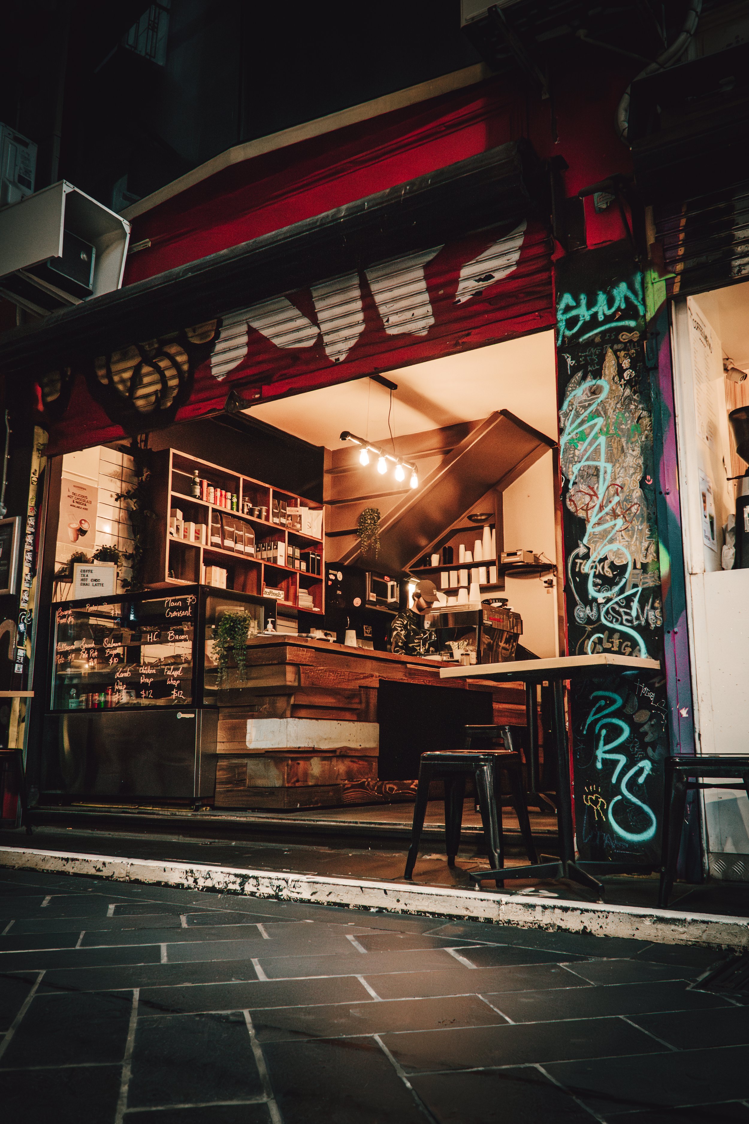 View of a small coffee shop with a wooden counter, shelves with coffee supplies, and a barista inside. Graffiti and chalkboard signs are visible outside. The shop has warm interior lighting and a chair in front.