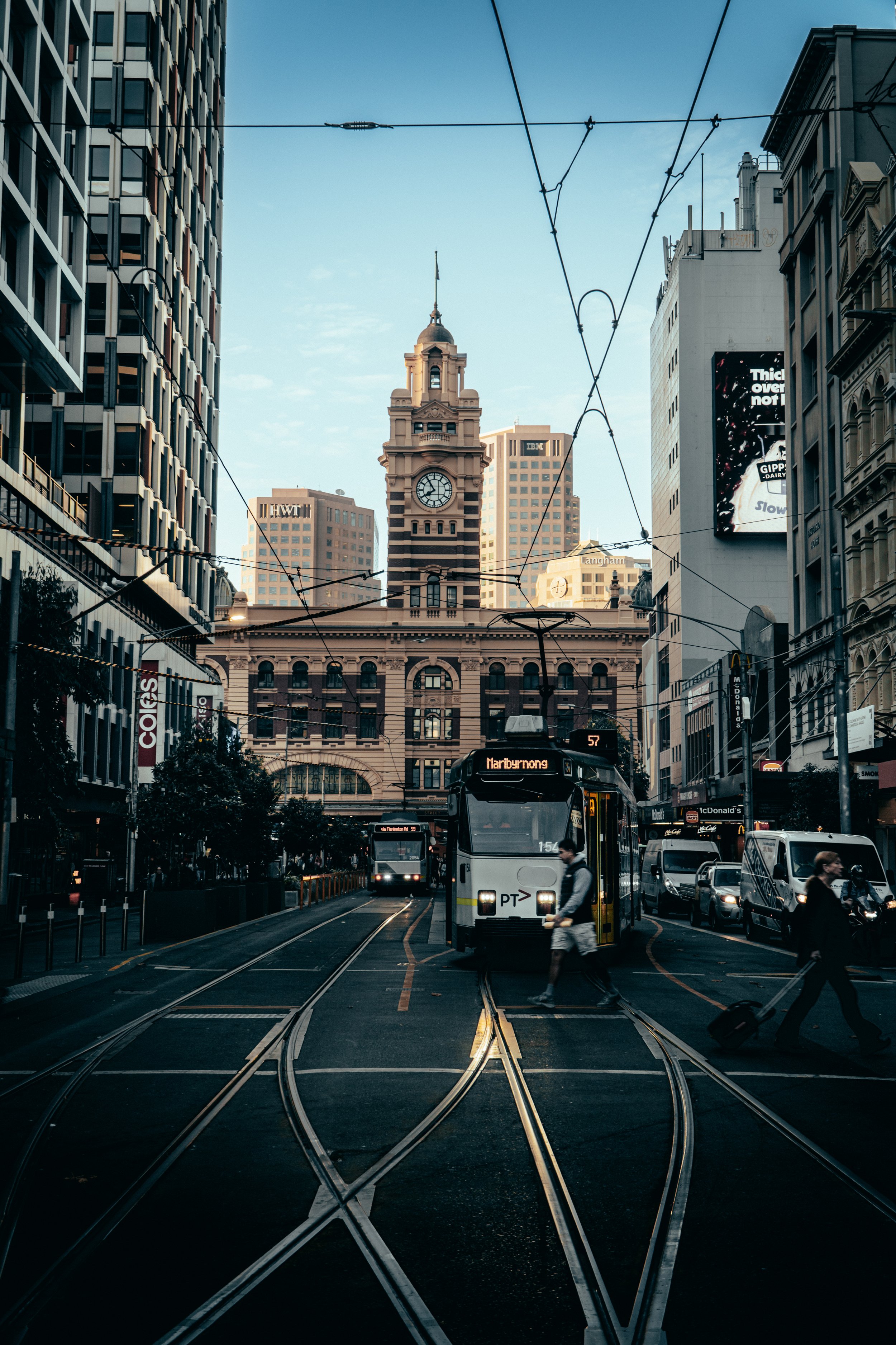 City street scene featuring a historic clock tower building in the background, modern skyscrapers on either side, trams on tracks, pedestrians crossing, and city traffic.