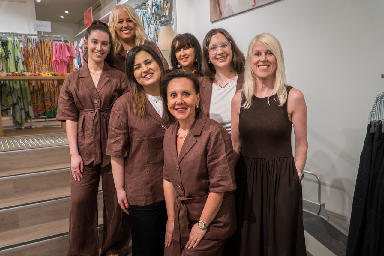 Group of seven women posing and smiling inside a clothing store with racks of colorful clothes in the background.